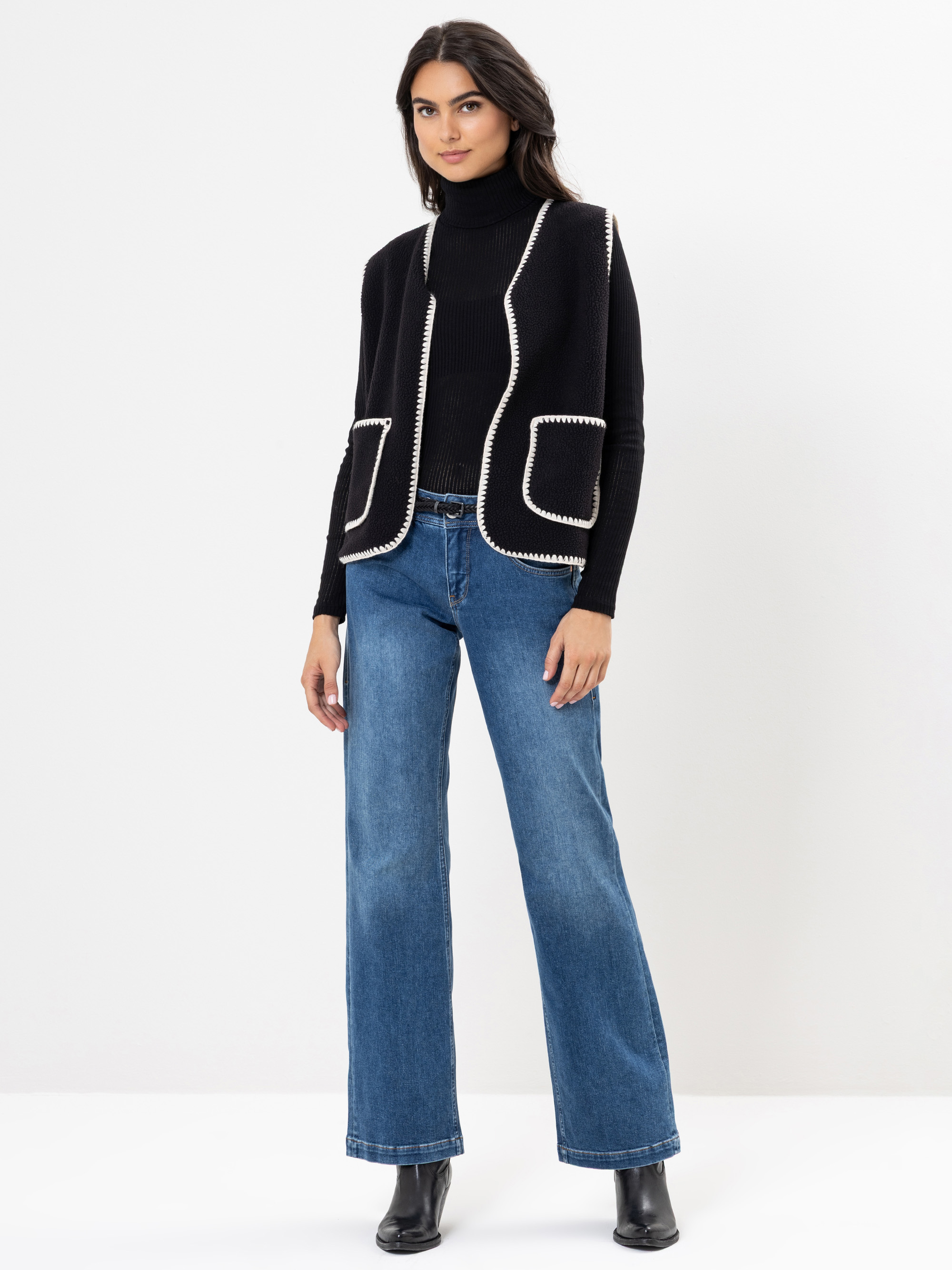 Eine Frau steht vor einem weißen Hintergrund und trägt einen schwarzen Rollkragenpullover, eine schwarze Weste mit weißem Saum, eine blaue Jeans mit weitem Bein und schwarze Ankle Boots. Sie hat langes dunkles Haar und einen neutralen Gesichtsausdruck.