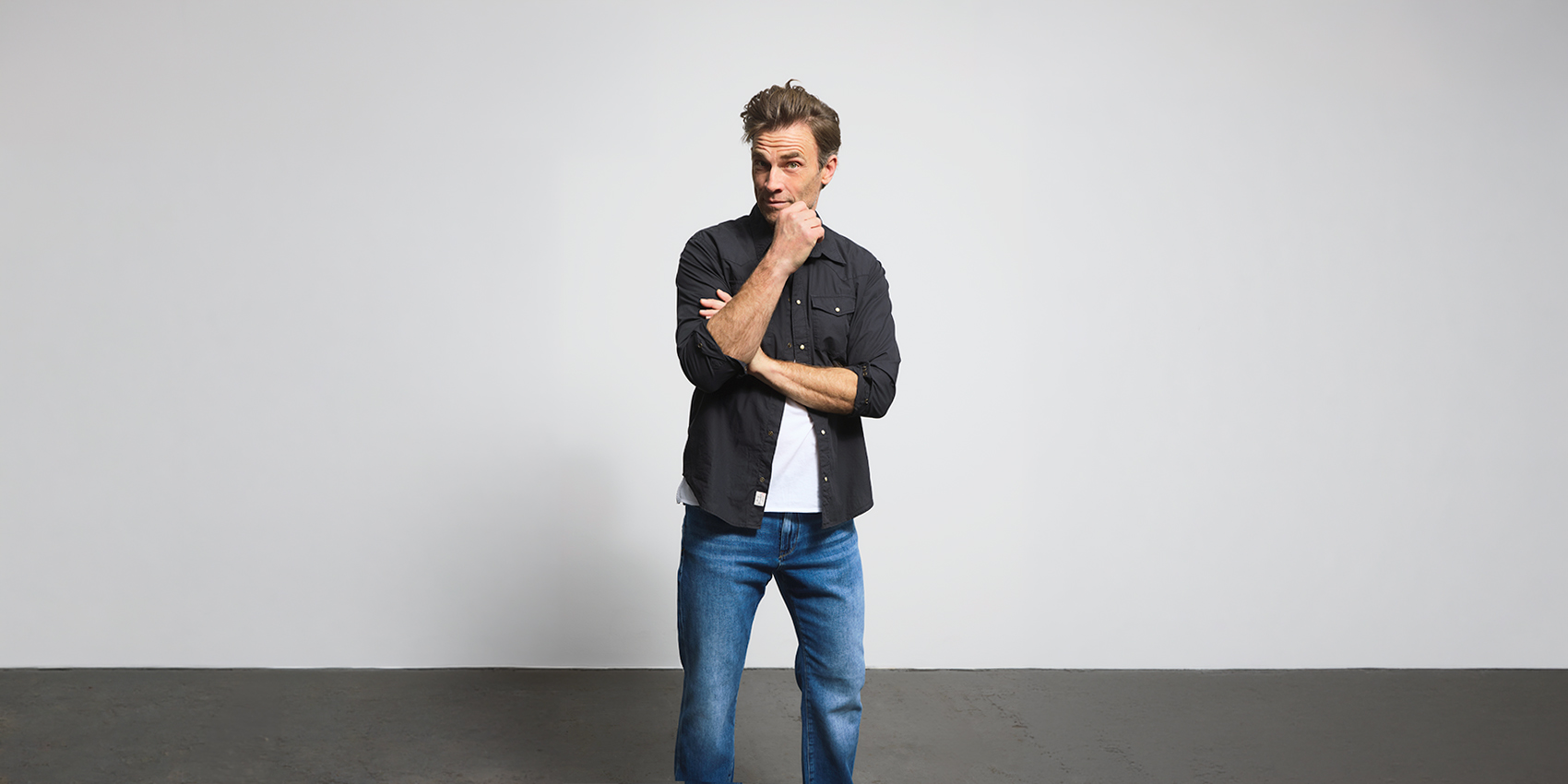 Ein Mann steht in einem minimalistisch eingerichteten Raum mit einer schlichten weißen Wand. Er trägt ein schwarzes Hemd über einem weißen T-Shirt und blaue Jeans. Er wirkt nachdenklich, stützt sein Kinn auf eine Hand und verschränkt die Arme.