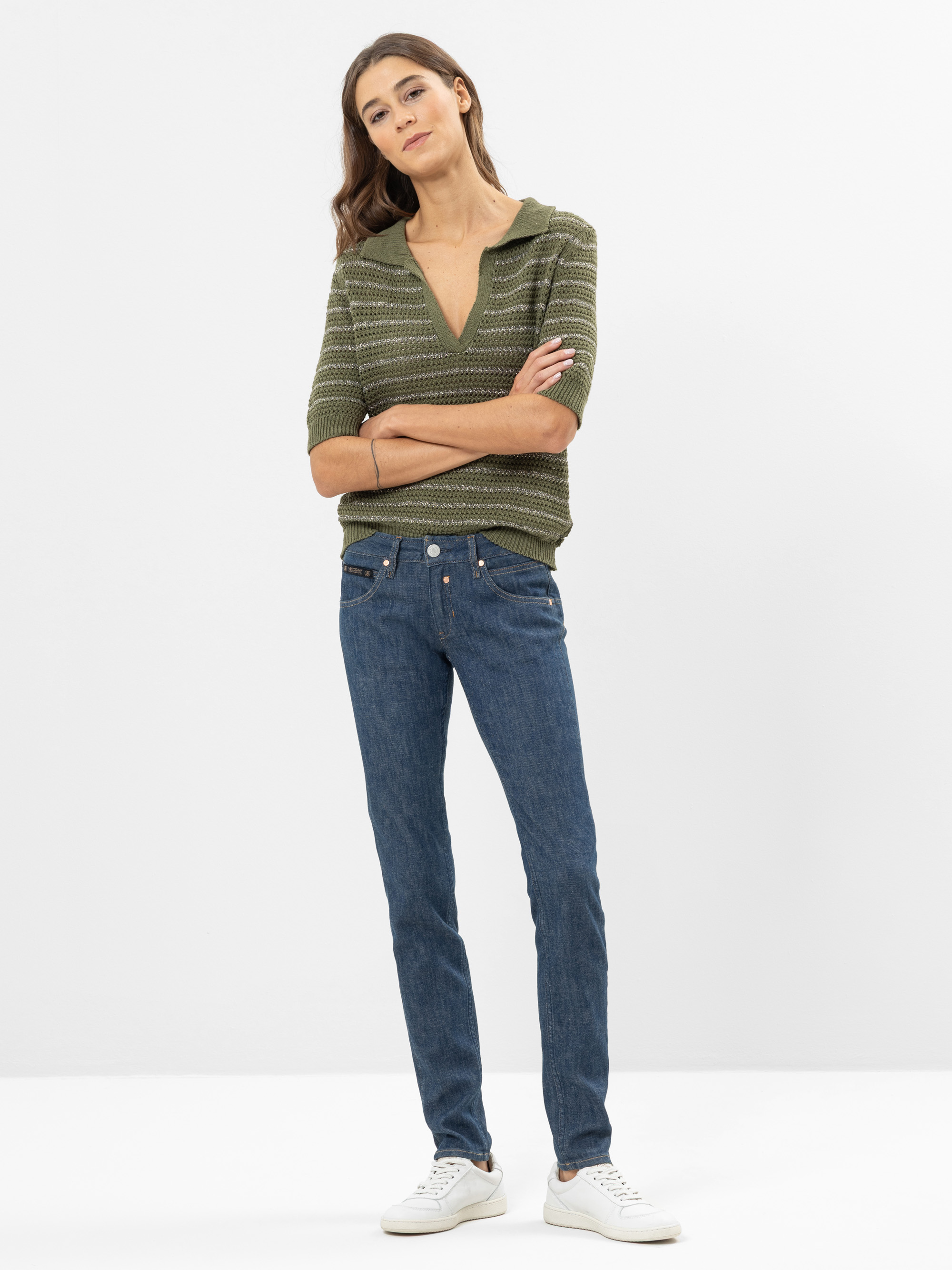 Eine Frau steht mit verschränkten Armen, einem grün gestreiften Pullover, blauen Jeans und weißen Turnschuhen vor einem schlichten weißen Hintergrund.