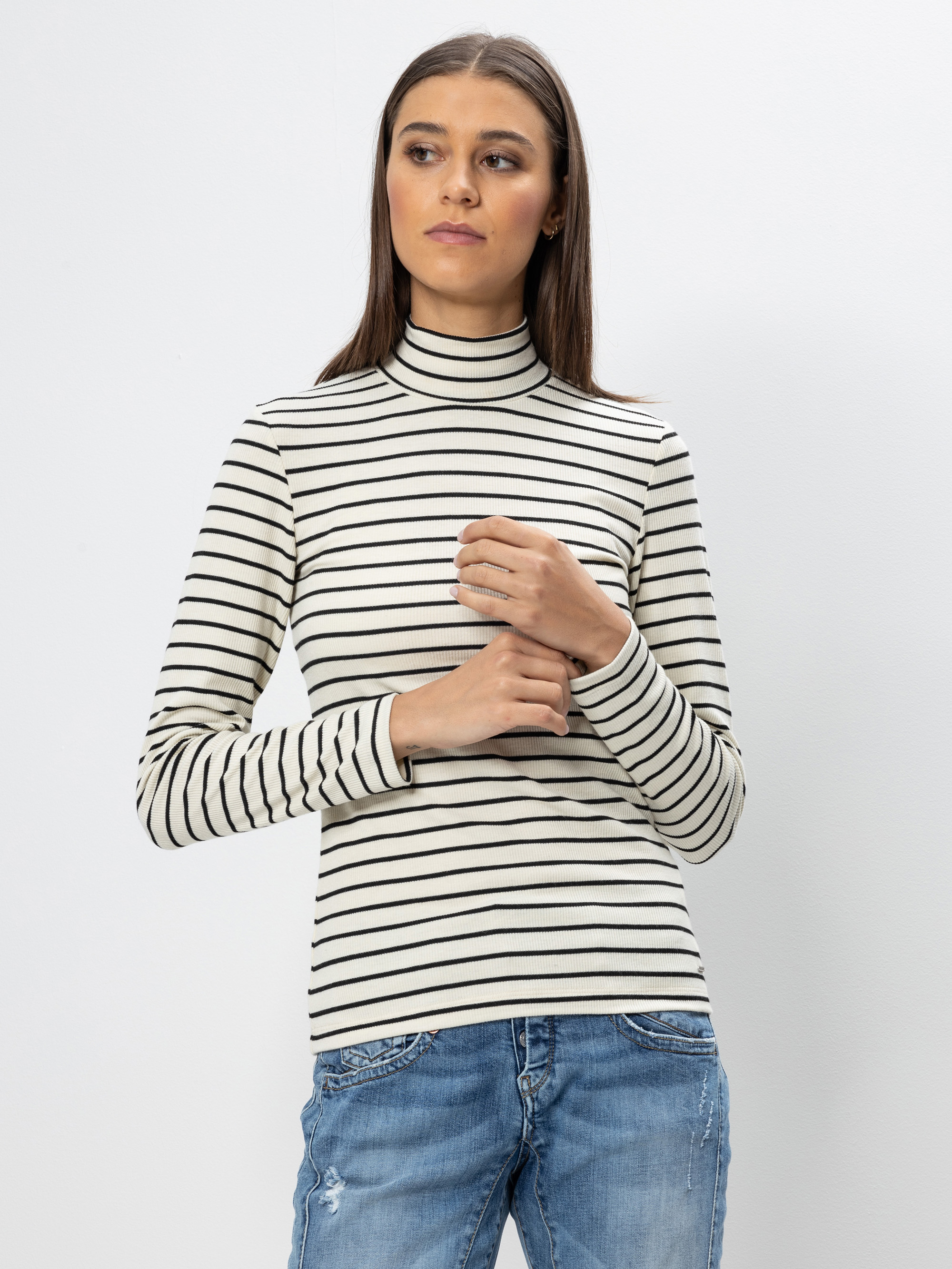 Eine Frau, die einen langärmeligen, schwarz-weiß gestreiften Rollkragenpullover und blaue Jeans trägt, steht vor einem schlichten weißen Hintergrund und blickt leicht zur Seite, während sie ihre Hände sanft verschränkt.