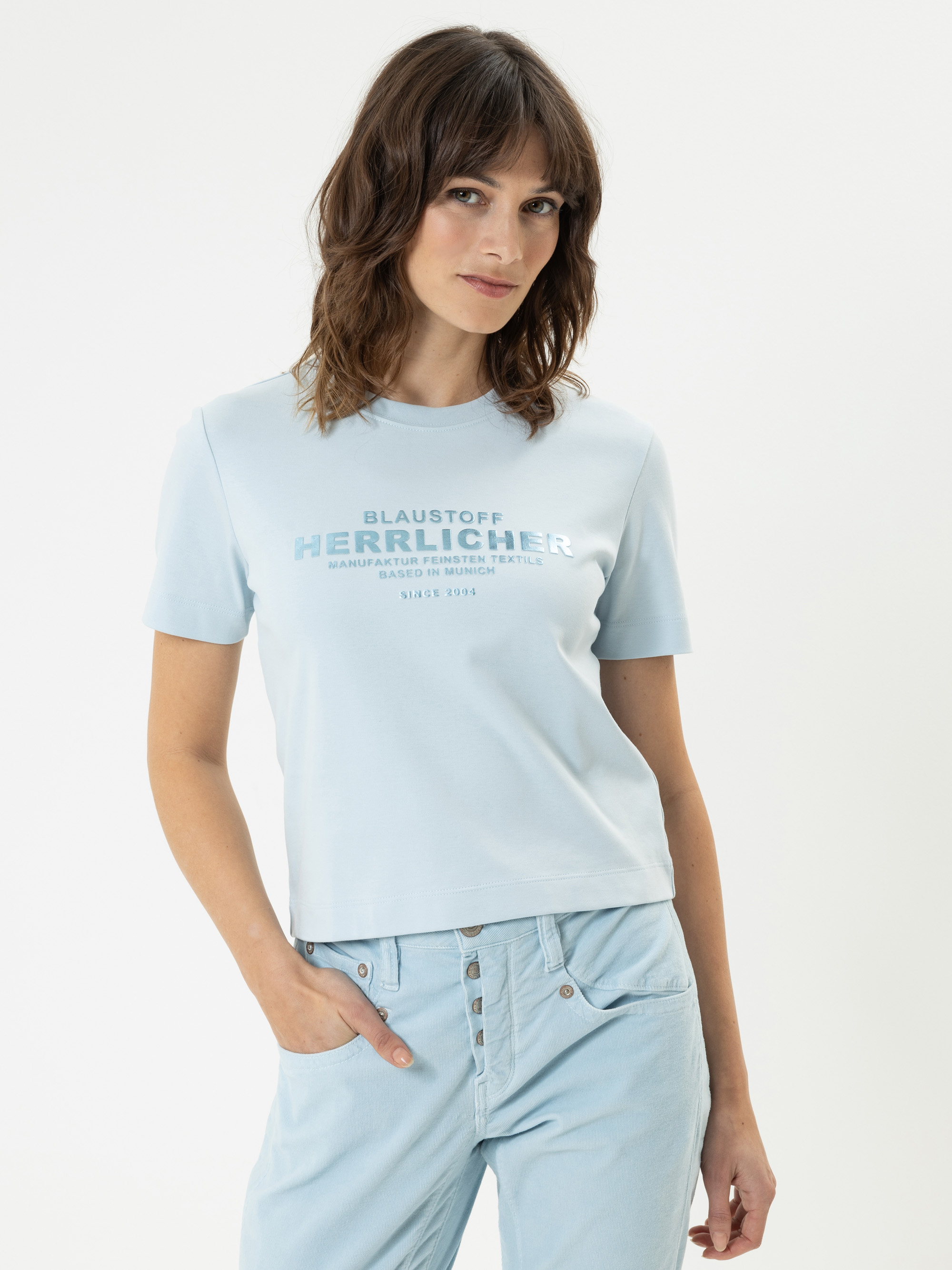 Eine Frau mit gewelltem braunem Haar trägt ein hellblaues T-Shirt mit Text auf der Vorderseite und eine hellblaue Hose. Sie steht vor einem weißen Hintergrund, hat eine Hand in der Tasche, schaut in die Kamera und lächelt sanft.