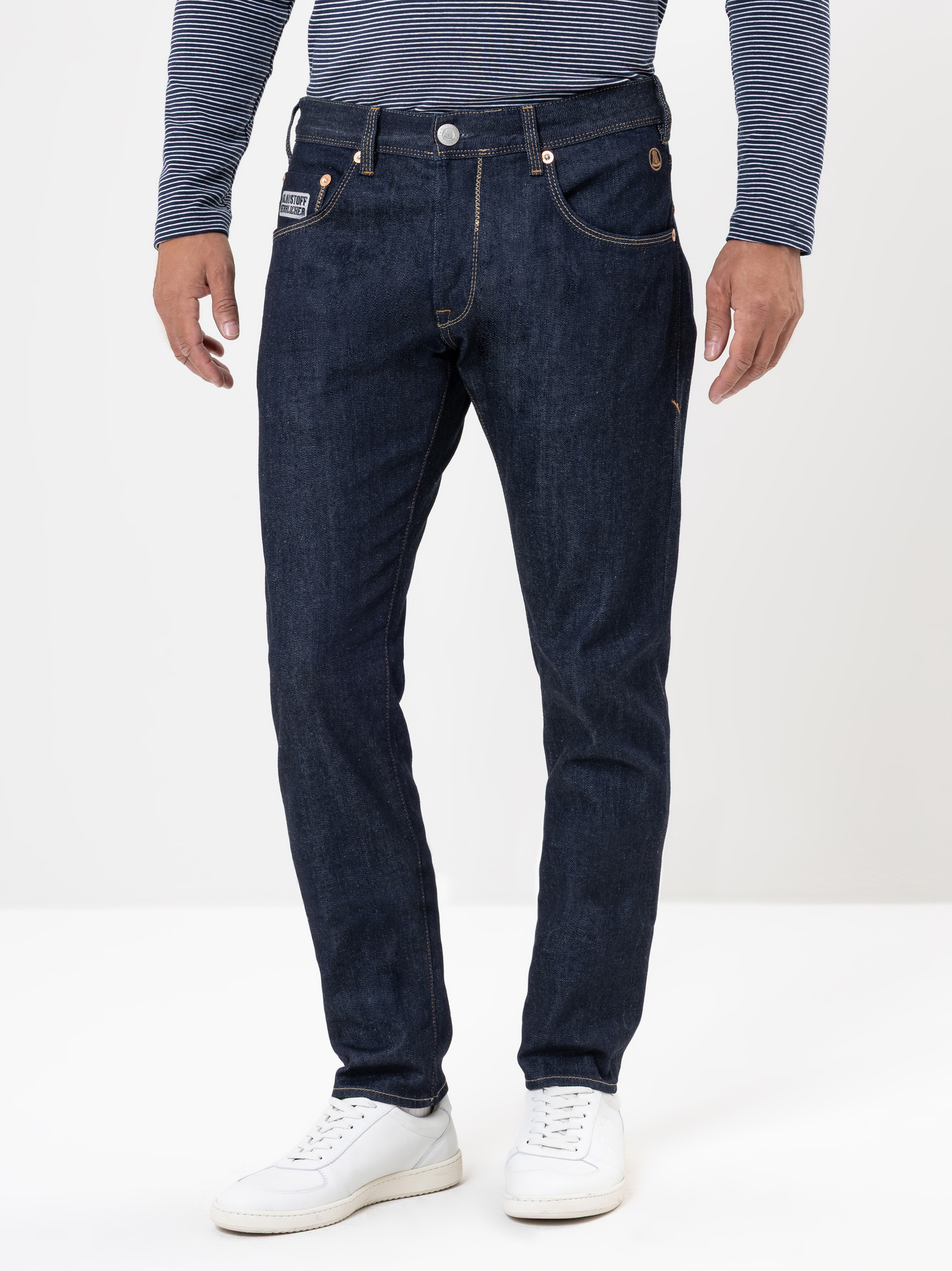 Eine Person steht vor einem weißen Hintergrund und trägt eine dunkelblaue Jeans, ein langärmeliges gestreiftes Hemd und weiße Turnschuhe. Nur der Körper von den Schultern abwärts ist sichtbar.
