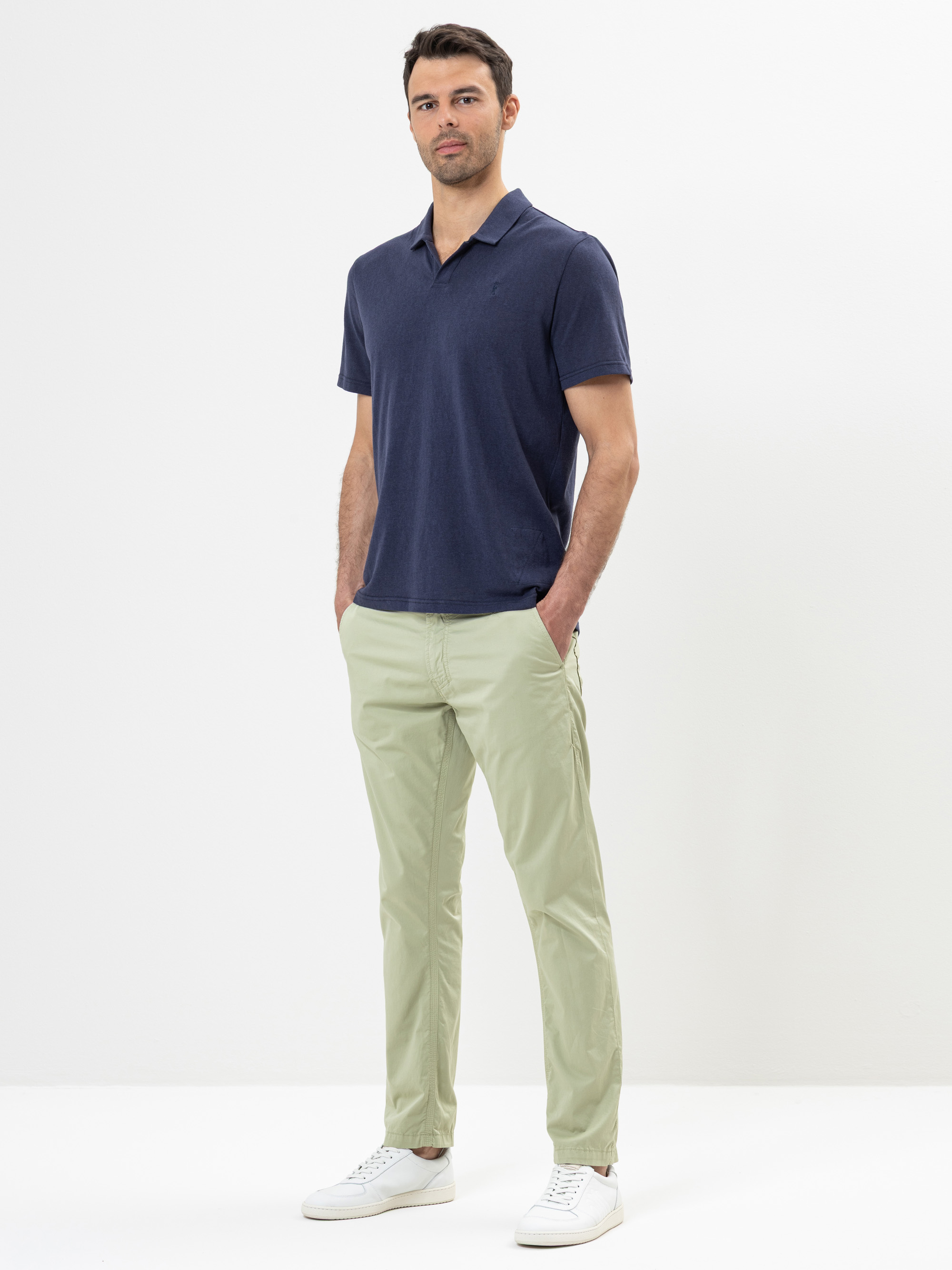 Ein Mann steht vor einem schlichten weißen Hintergrund und trägt ein marineblaues, kurzärmeliges Poloshirt, eine hellgrüne Hose und weiße Turnschuhe. Er hat kurzes dunkles Haar und seine Hände stecken in den Taschen.