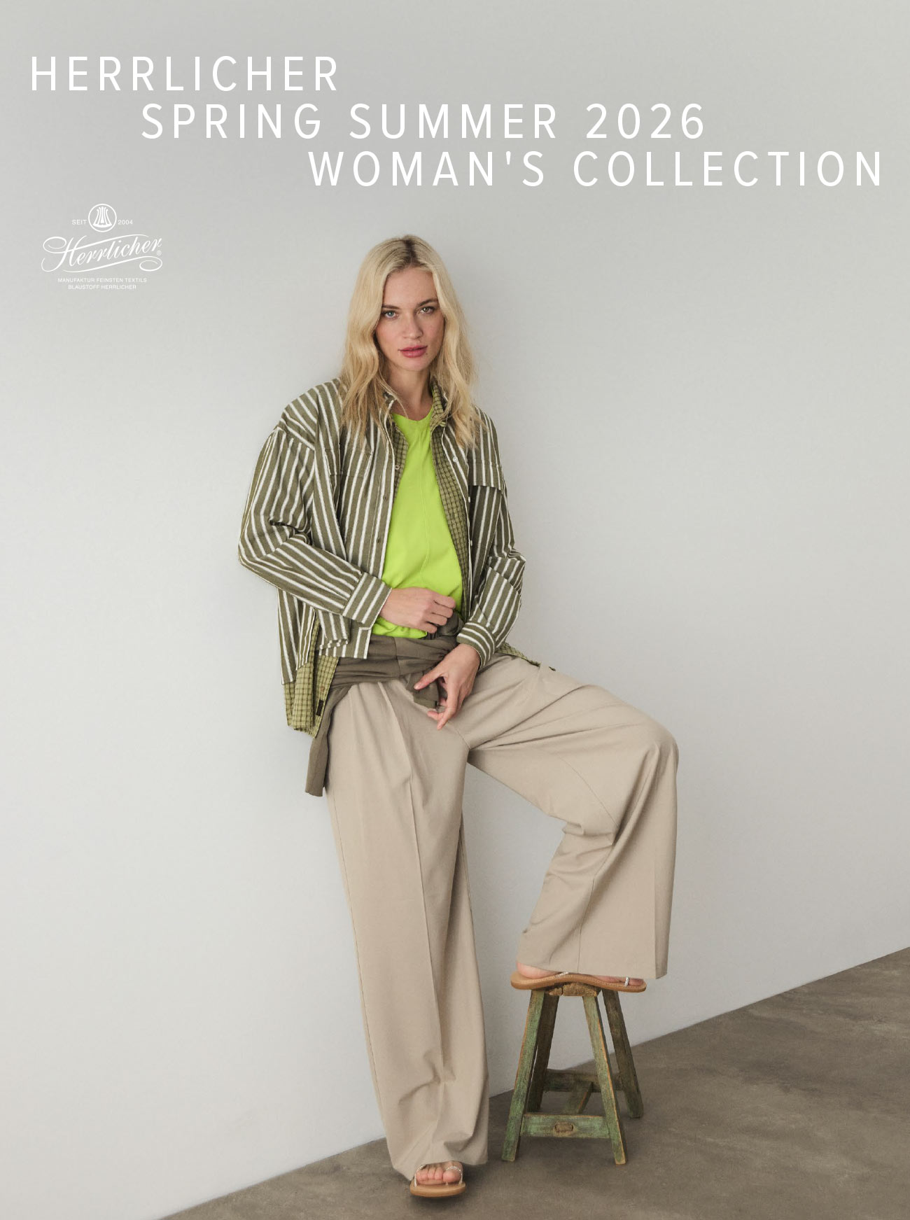 Eine Frau in einem grünen Oberteil, einer gestreiften Jacke und einer weiten beigen Hose posiert auf einem Holzschemel vor einem schlichten Hintergrund. Der Text lautet: HERRLICHER FRÜHLING SOMMER 2026 DAMENKOLLEKTION.