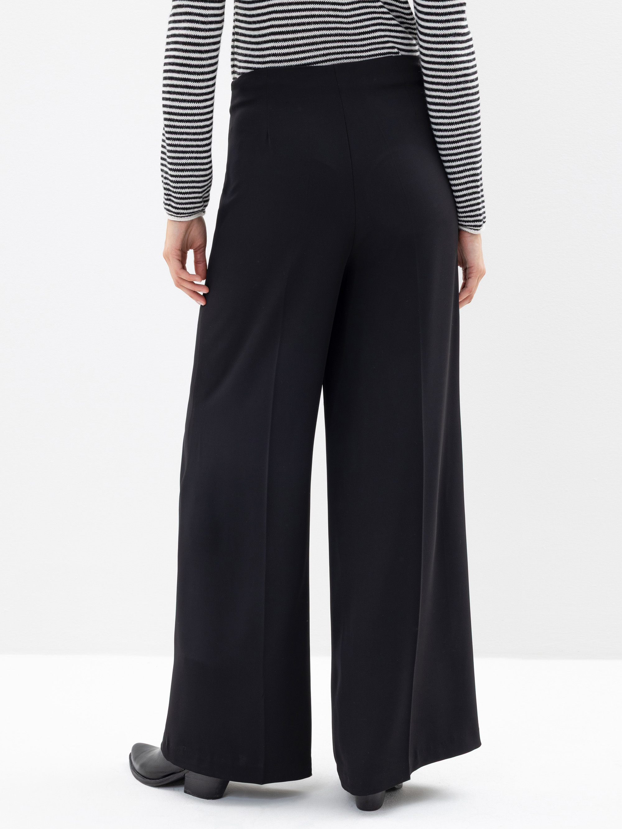 Eine Person, die ein schwarz-weiß gestreiftes langärmeliges Oberteil und eine locker sitzende schwarze Hose mit weitem Bein trägt, von hinten gesehen, steht vor einem schlichten weißen Hintergrund.