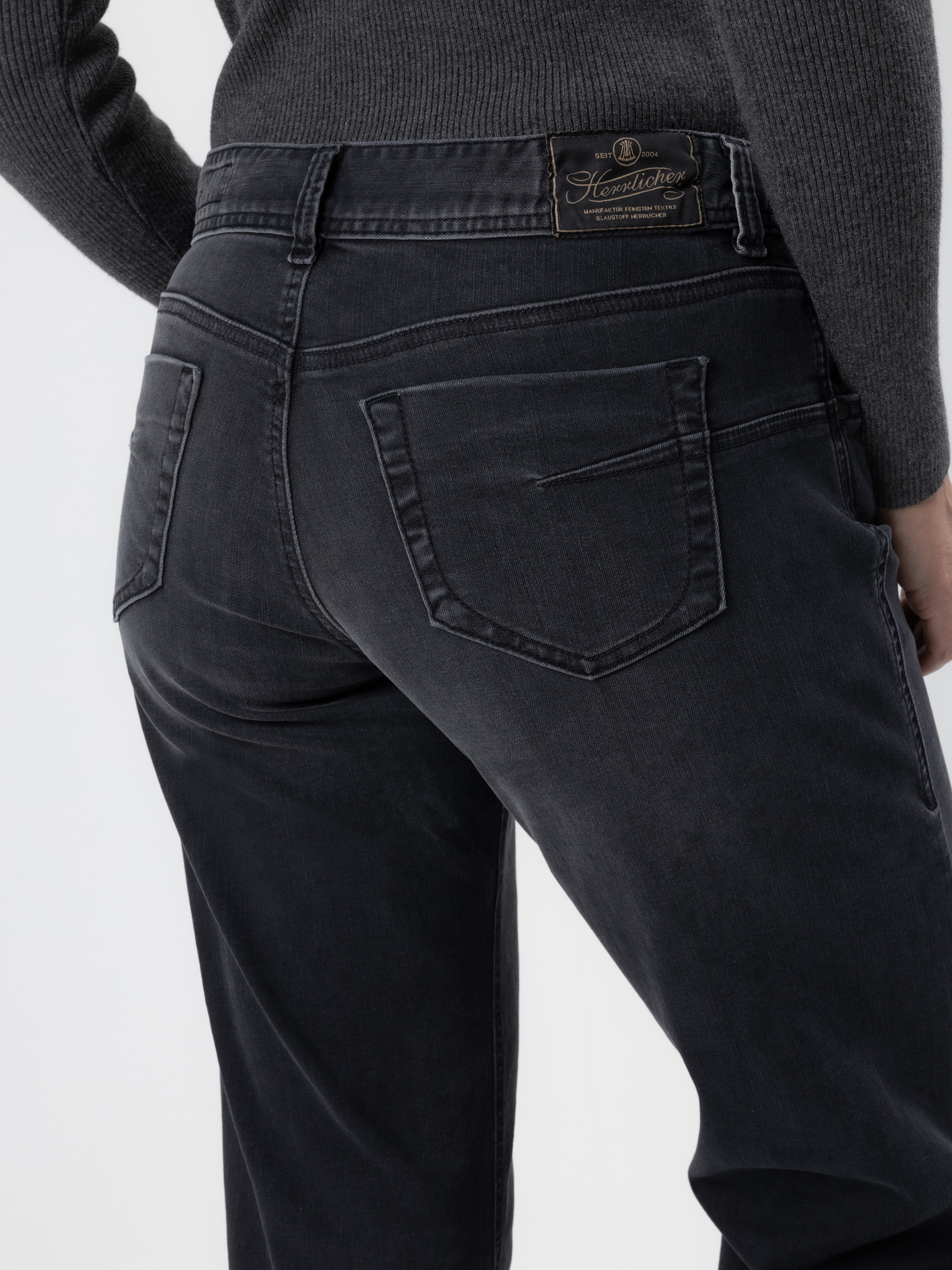 Nahaufnahme einer Person, die eine dunkelgraue Jeans und einen grauen gerippten Pullover trägt, wobei die Gesäßtasche und das Markenetikett am Bund zu sehen sind. Die Person steht vor einem schlichten hellen Hintergrund.