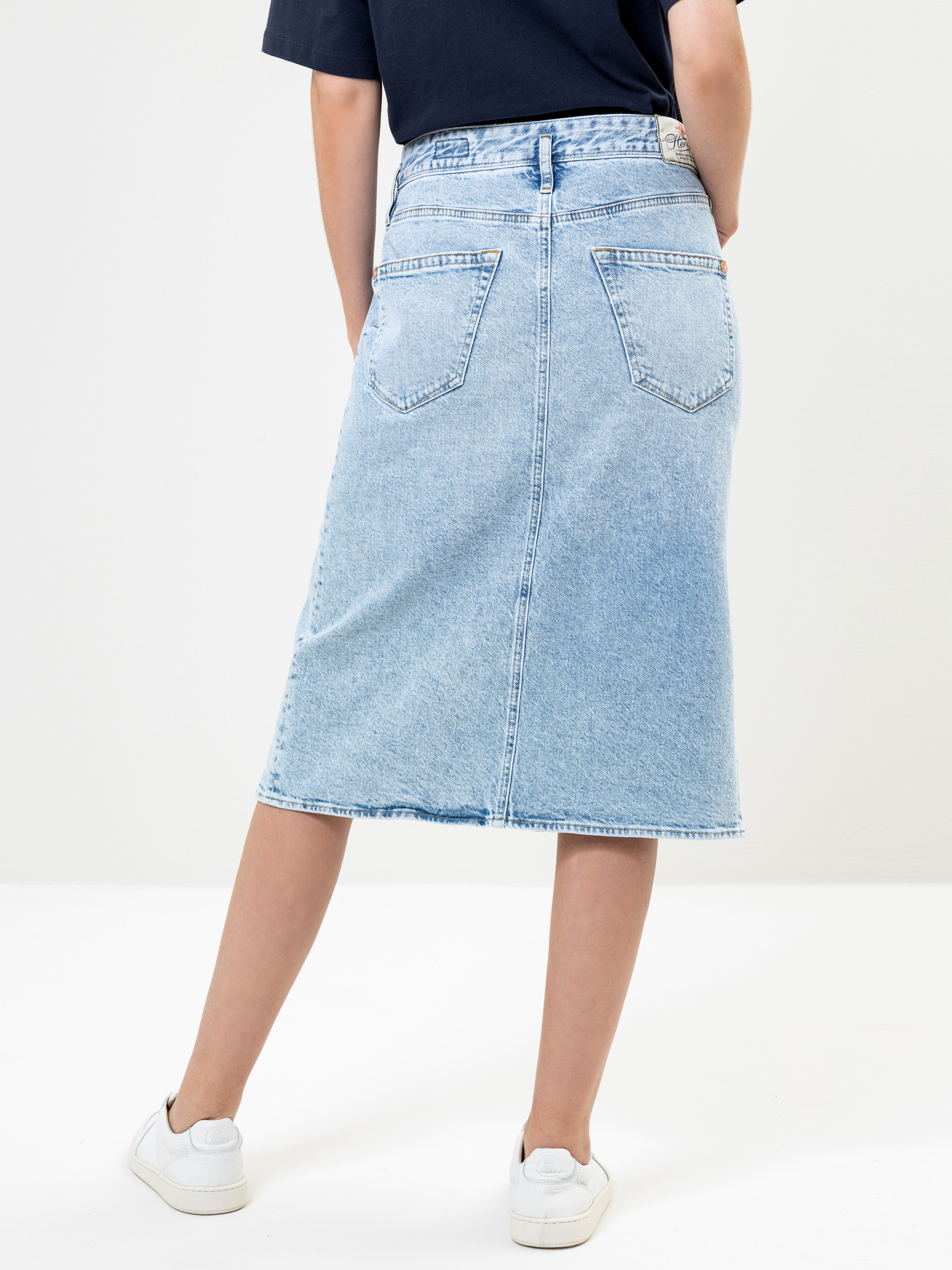Eine Person, die einen hellblauen Jeans-Midirock, ein dunkles Top und weiße Turnschuhe trägt, steht mit dem Rücken zur Kamera vor einem schlichten Hintergrund.