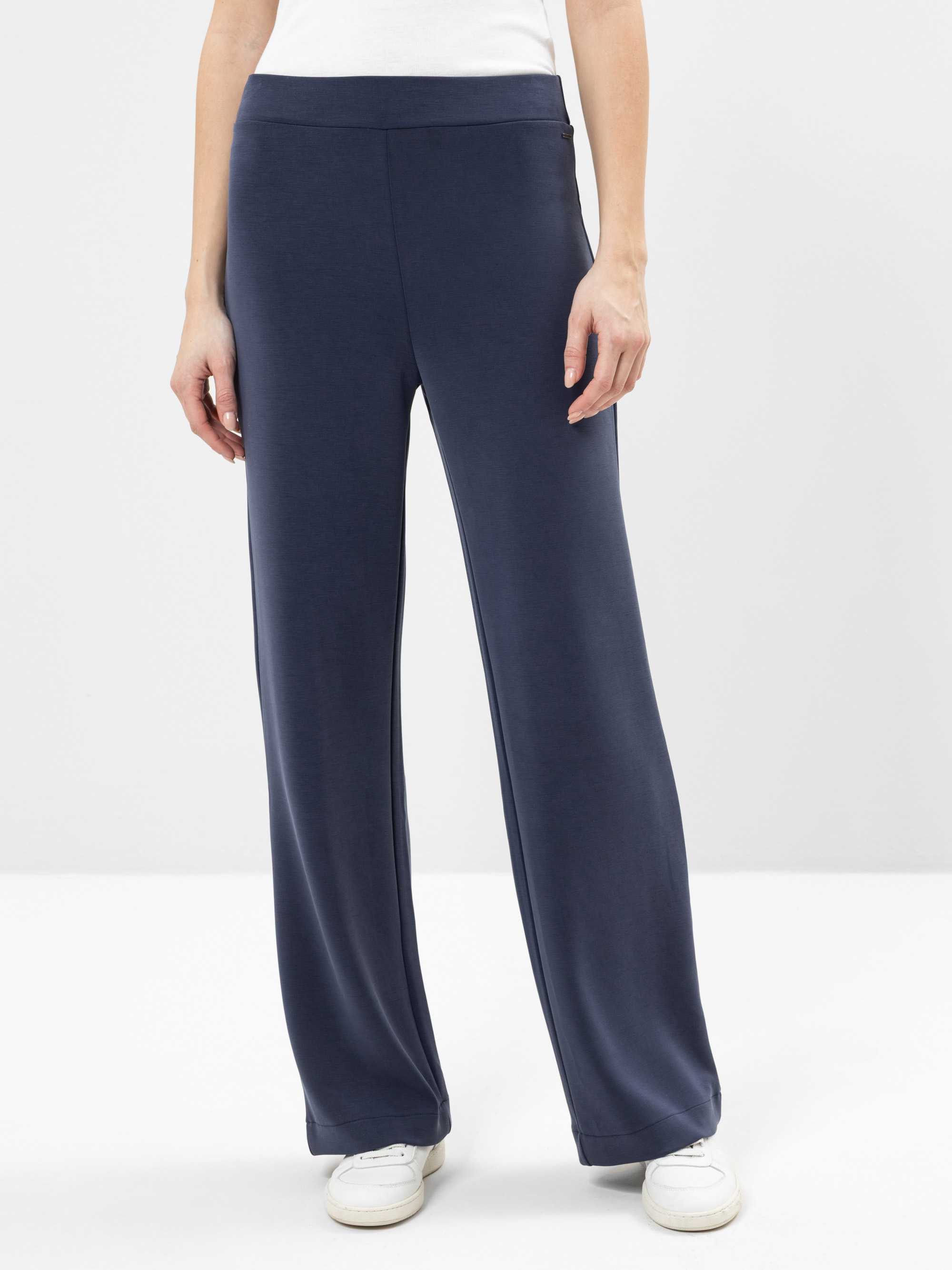 Eine Person, die eine locker sitzende marineblaue Hose mit weitem Bein, ein weißes Oberteil und weiße Turnschuhe trägt, steht vor einem schlichten weißen Hintergrund. Das Bild ist von den Schultern abwärts beschnitten.
