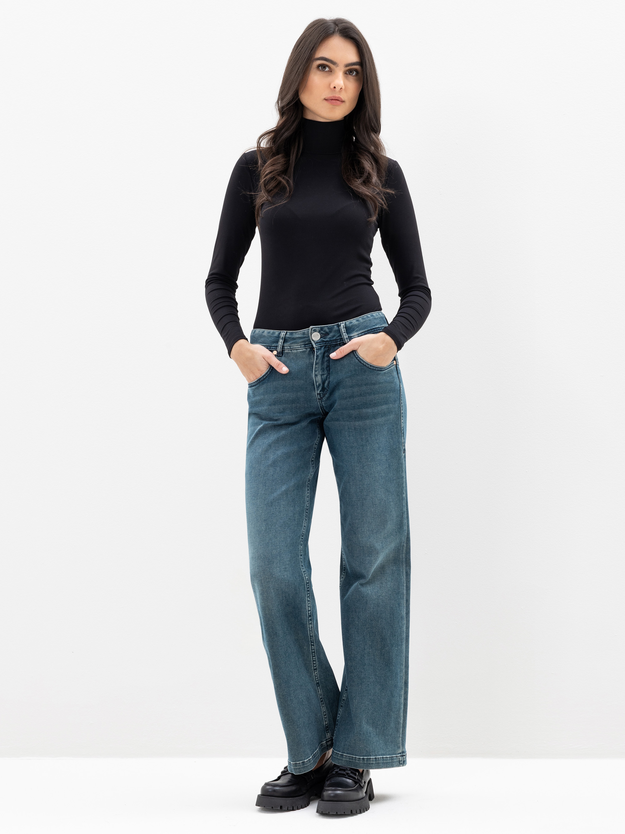 Eine Frau steht vor einem schlichten weißen Hintergrund und trägt einen schwarzen Rollkragenpullover, blaue Jeans mit weitem Bein und schwarze Schuhe. Sie hat die Hände in den Taschen und einen neutralen Gesichtsausdruck.