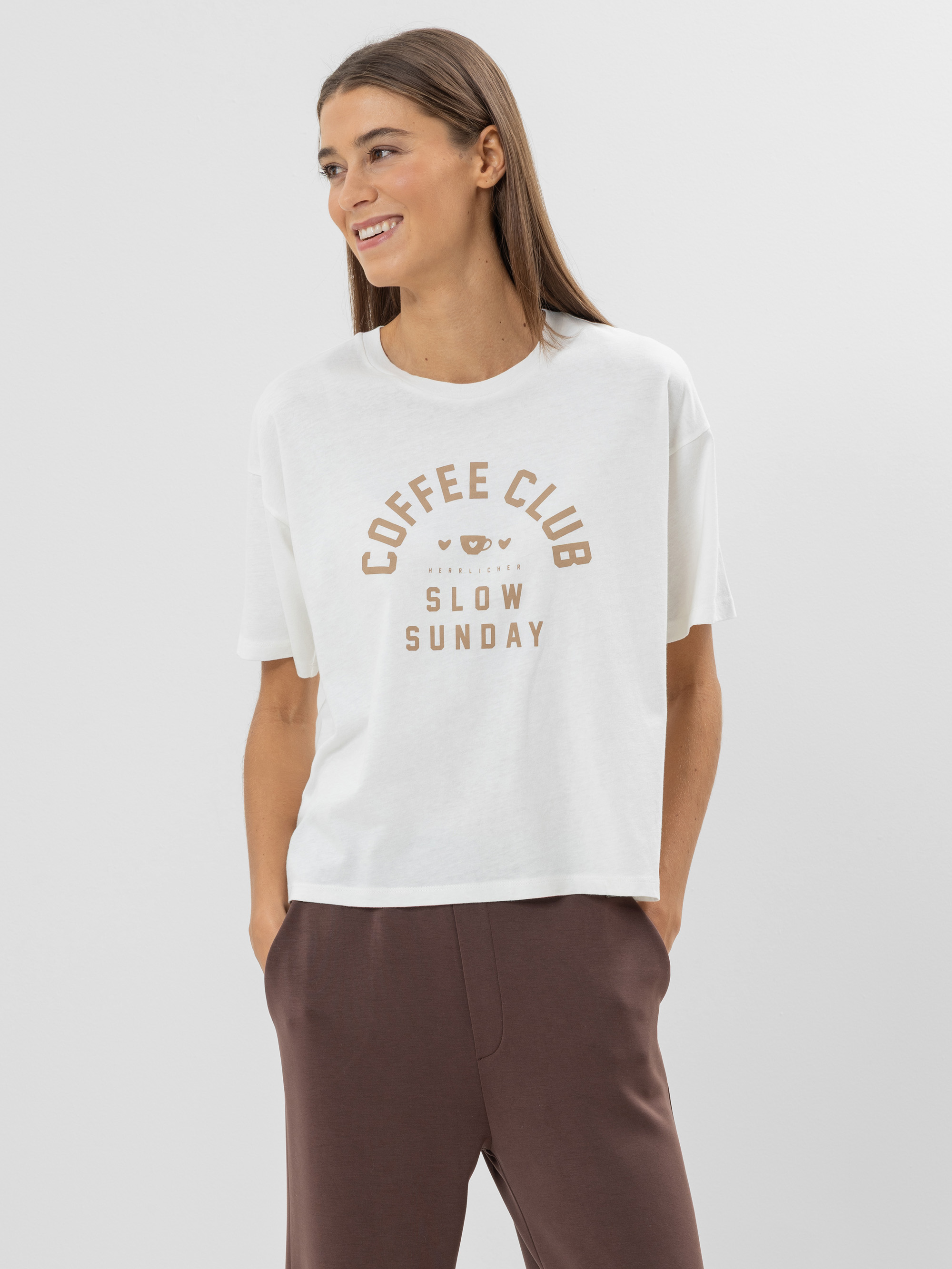 Frau mit langen braunen Haaren, die ein weißes T-Shirt trägt, auf dem in braunen Buchstaben "Coffee Club Slow Sunday" steht, und eine braune Hose, die vor einem schlichten hellen Hintergrund steht und zur Seite lächelt.