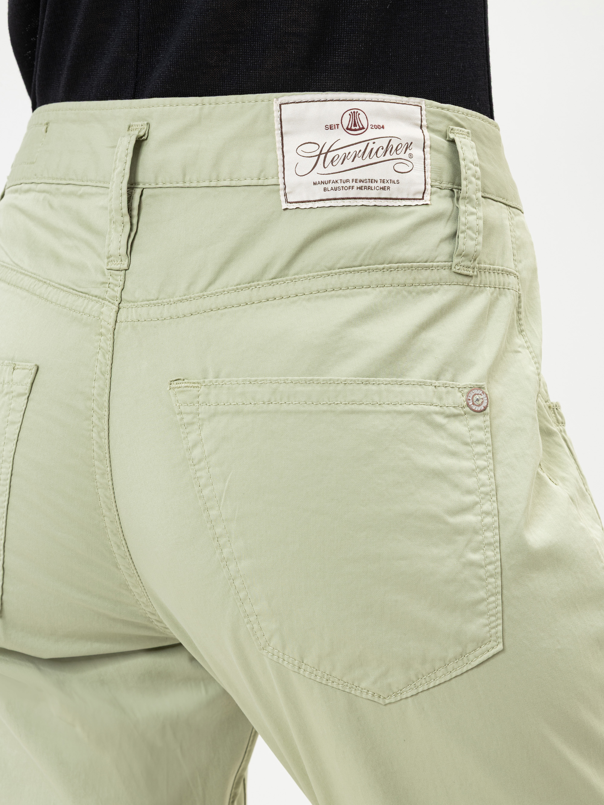Nahaufnahme der Rückseite einer hellgrünen Hose mit zwei Gesäßtaschen und einem weißen Herrlicher-Markenetikett, das über der rechten Tasche aufgenäht ist. Eine Person trägt die Hose mit einem teilweise sichtbaren schwarzen Oberteil.