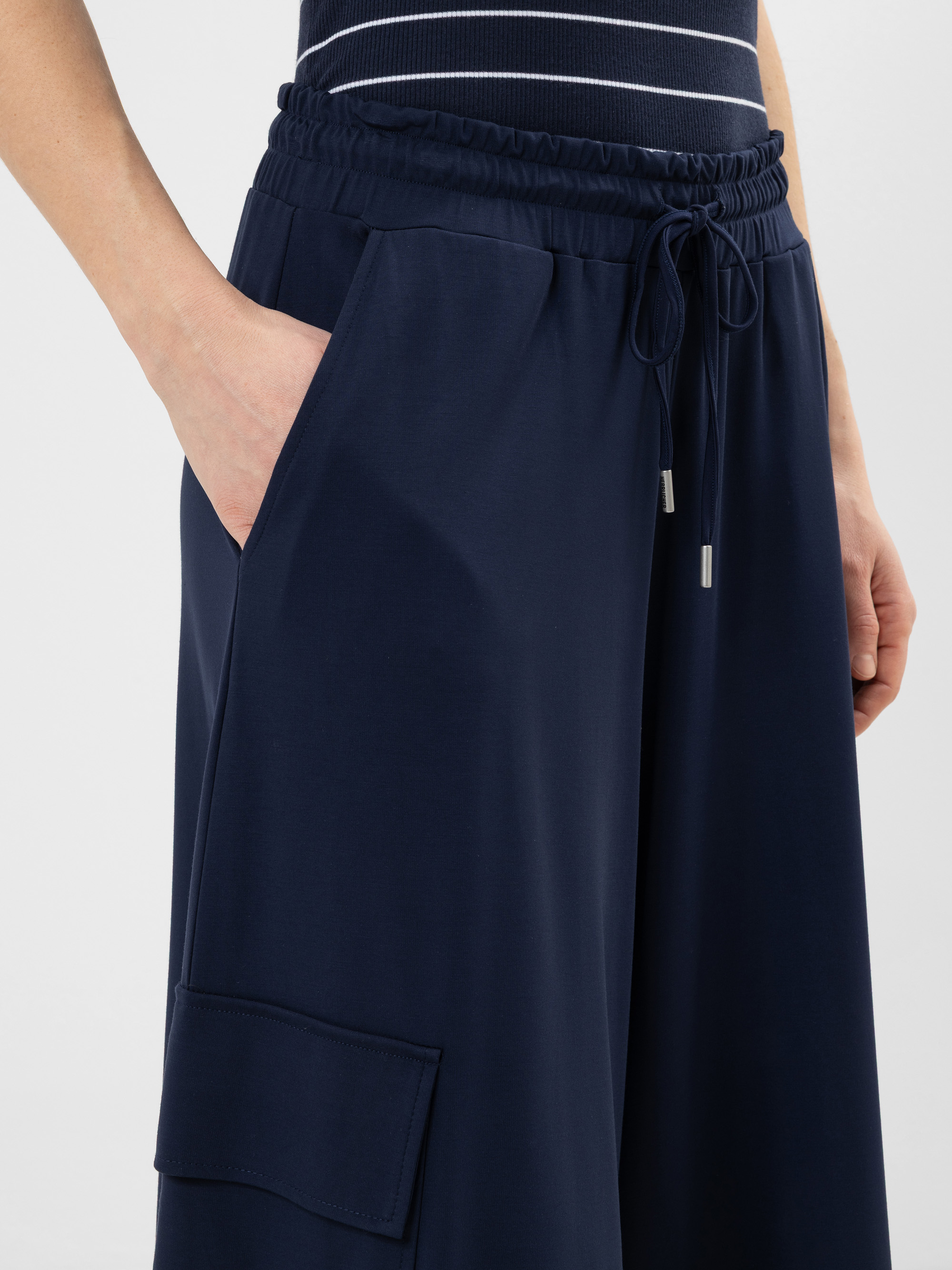 Eine Person trägt eine locker sitzende marineblaue Hose mit einem elastischen Kordelzug in der Taille und einer Tasche an der Seite, gepaart mit einem marineblau-weiß gestreiften Oberteil. Eine Hand befindet sich in einer Tasche.
