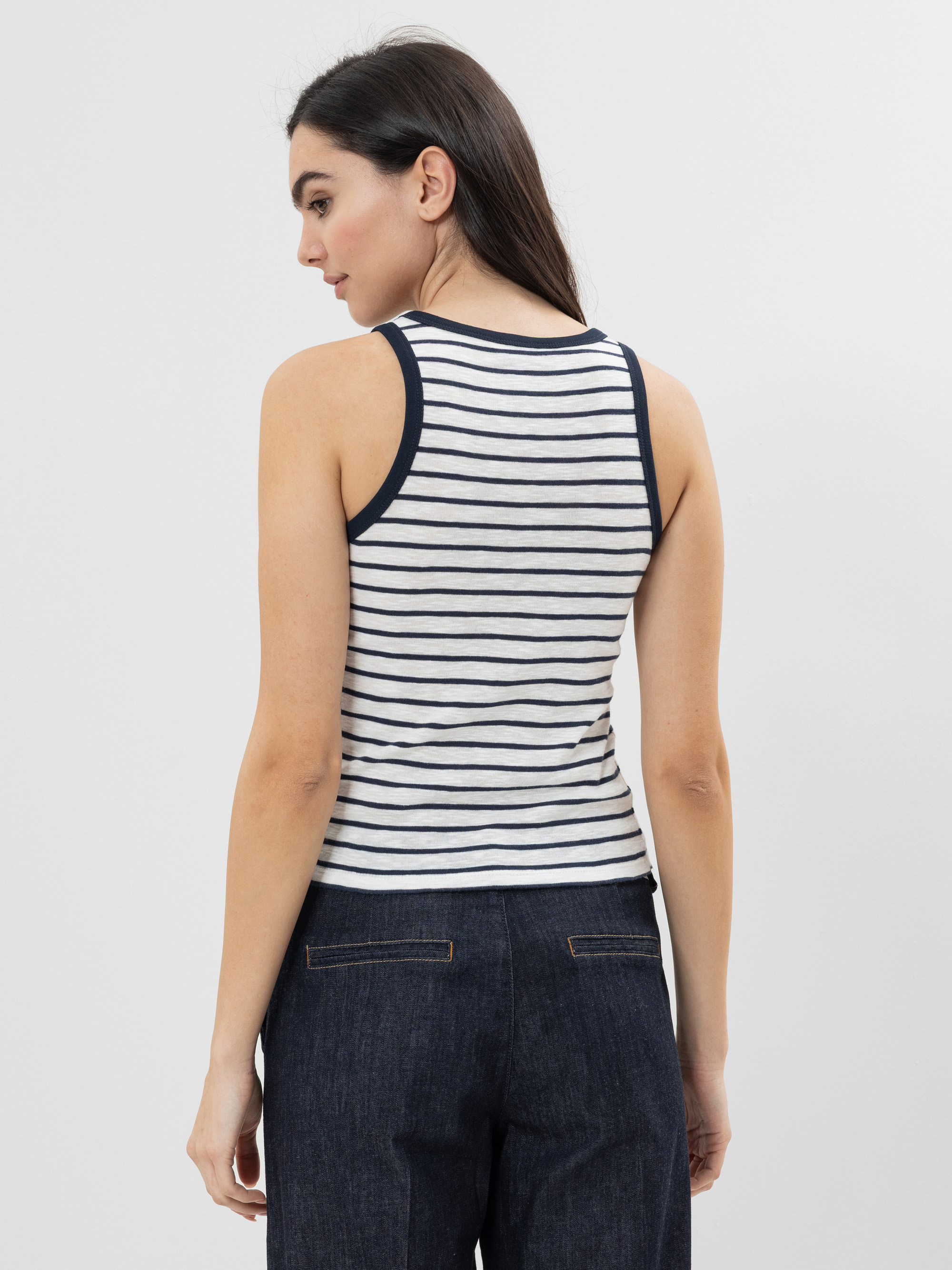Frau, von hinten gesehen, trägt ein ärmelloses, weißes Tanktop mit schwarzen Querstreifen und dunkelblaue Jeans. Sie hat langes, dunkles Haar und steht vor einem schlichten, hellen Hintergrund.