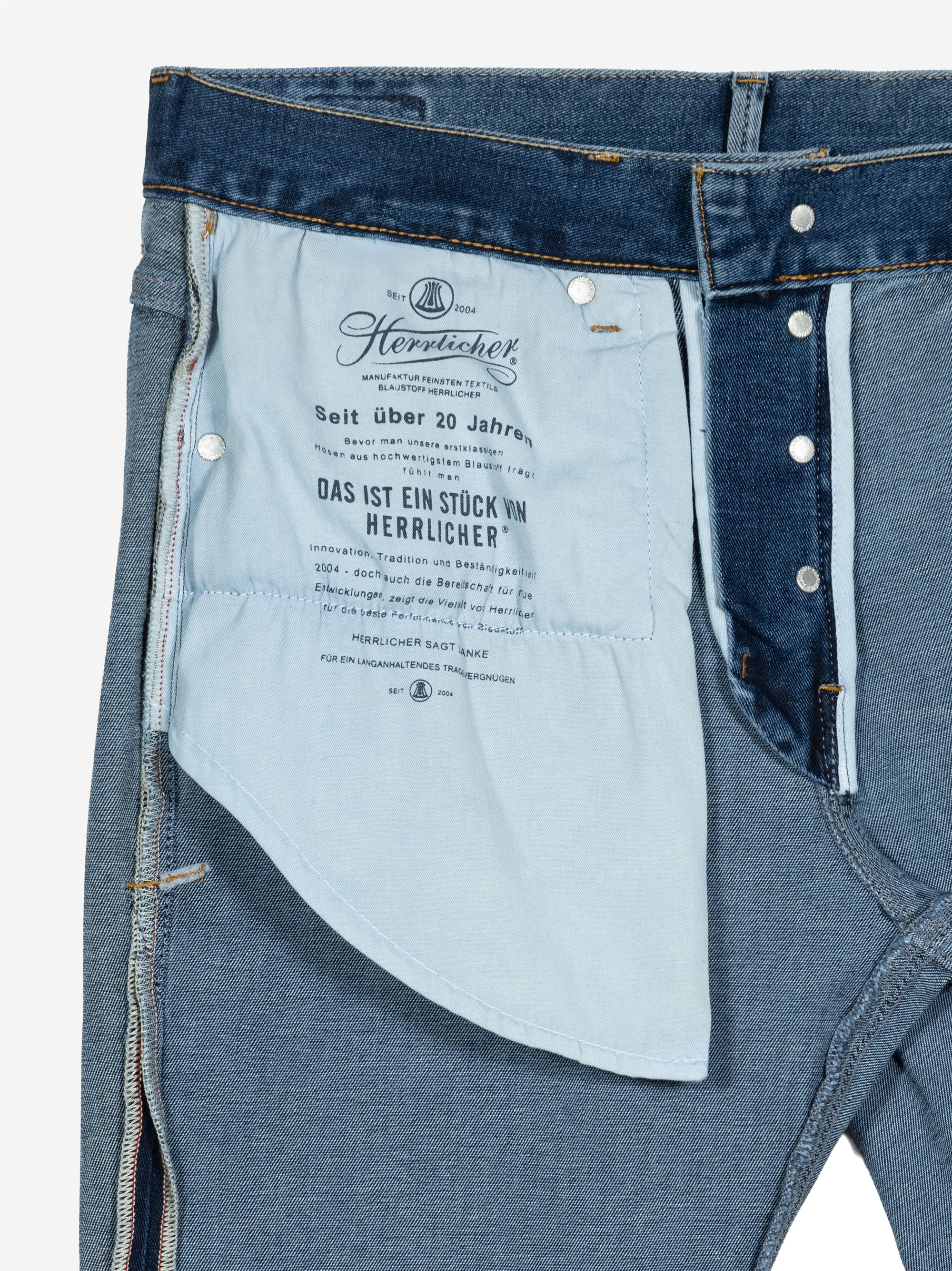 Nahaufnahme einer blauen Jeans, bei der die Vorderseite aufgeknöpft und das Taschenfutter freigelegt ist und ein bedrucktes Etikett mit Markenzeichen und Text in deutscher Sprache zu sehen ist.