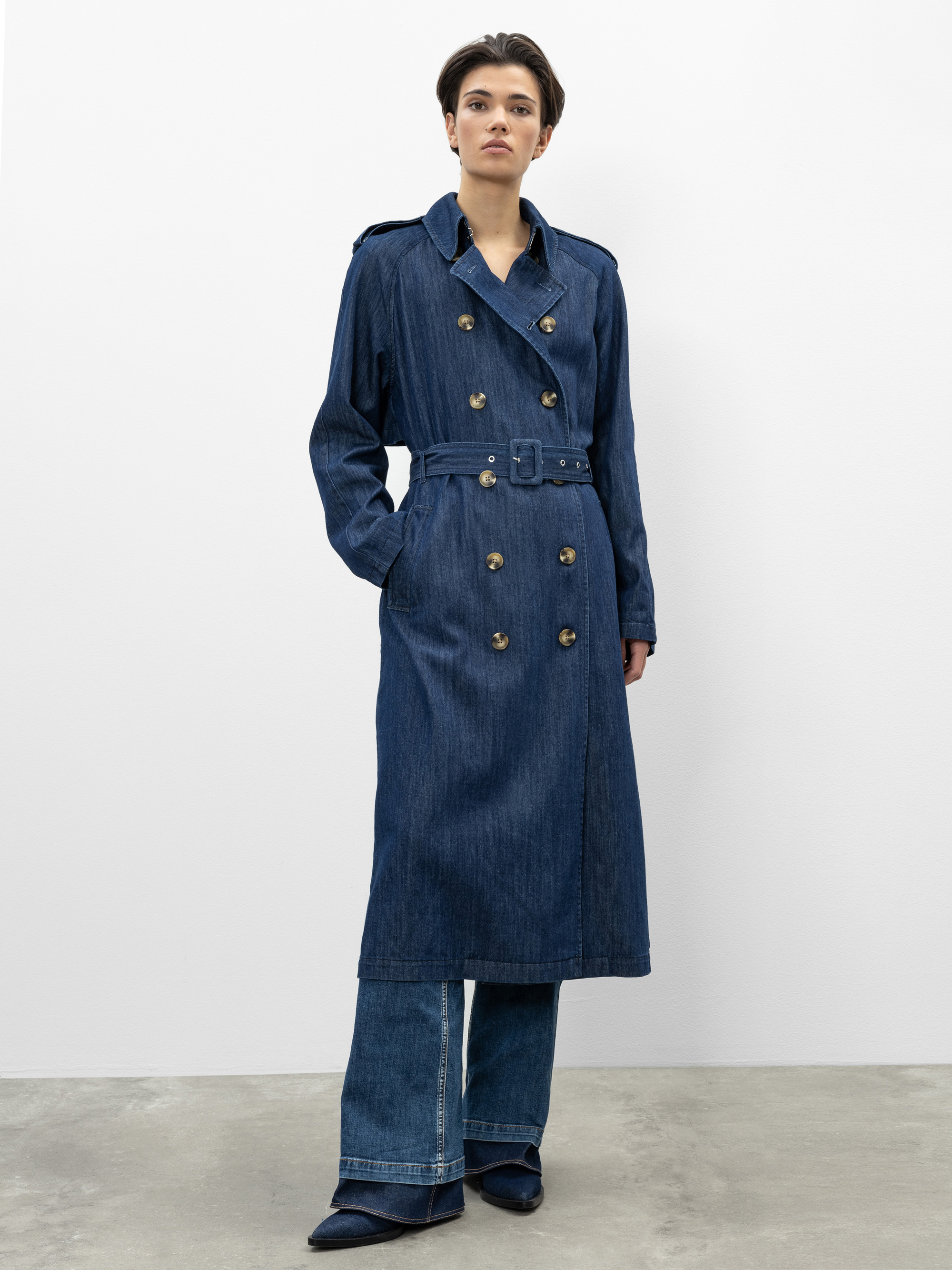 Eine Person steht vor einer schlichten weißen Wand und trägt einen langen, gegürteten Trenchcoat aus Denim über einer Jeans mit weitem Bein und dunklen spitzen Schuhen. Sie hat kurzes Haar und einen neutralen Gesichtsausdruck, die Hände stecken in de