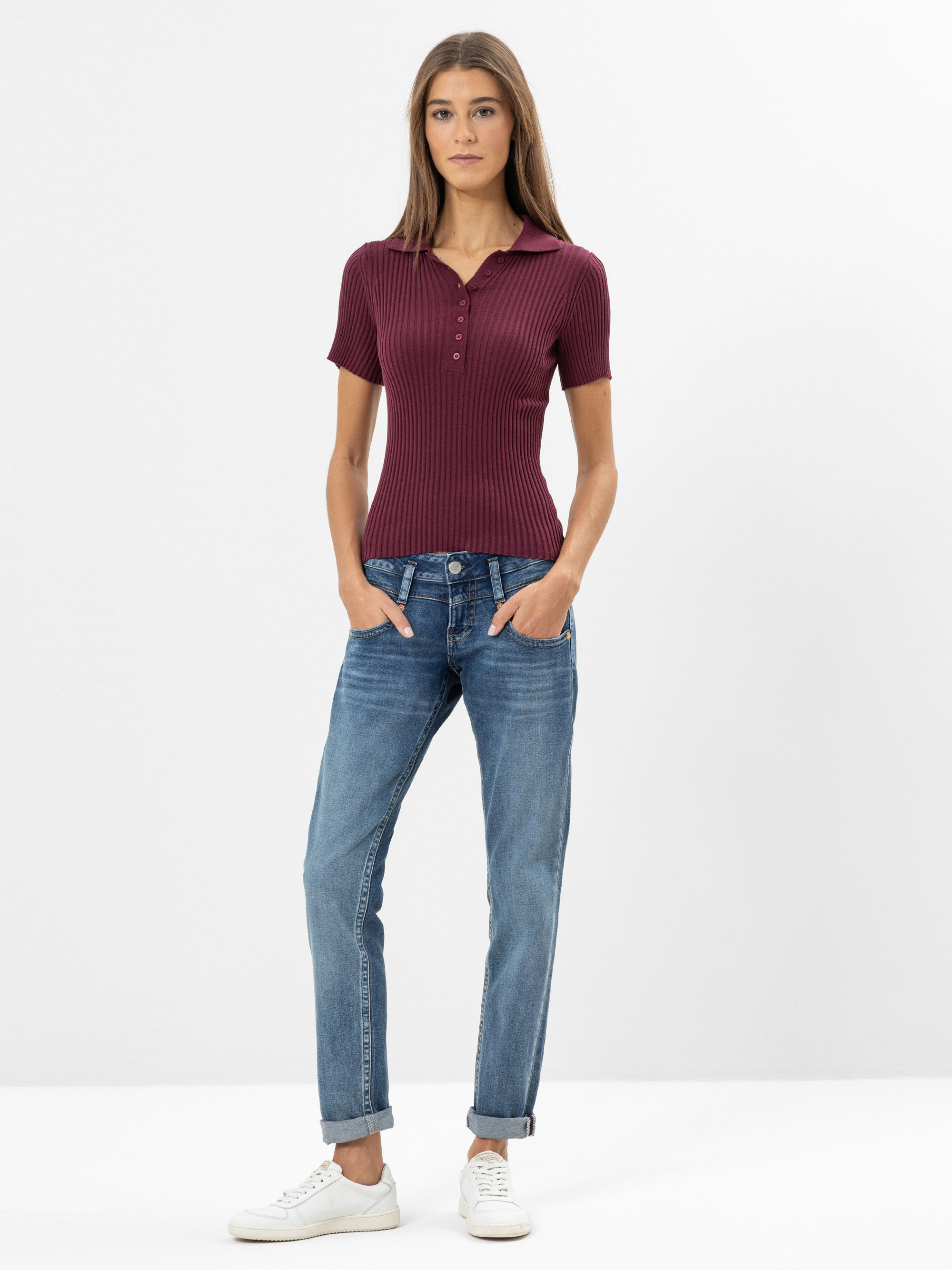 Eine Frau steht mit dem Gesicht nach vorne, die Hände in den Taschen ihrer blauen Jeans, trägt ein kurzärmeliges, geripptes bordeauxfarbenes Poloshirt und weiße Turnschuhe vor einem schlichten weißen Hintergrund.