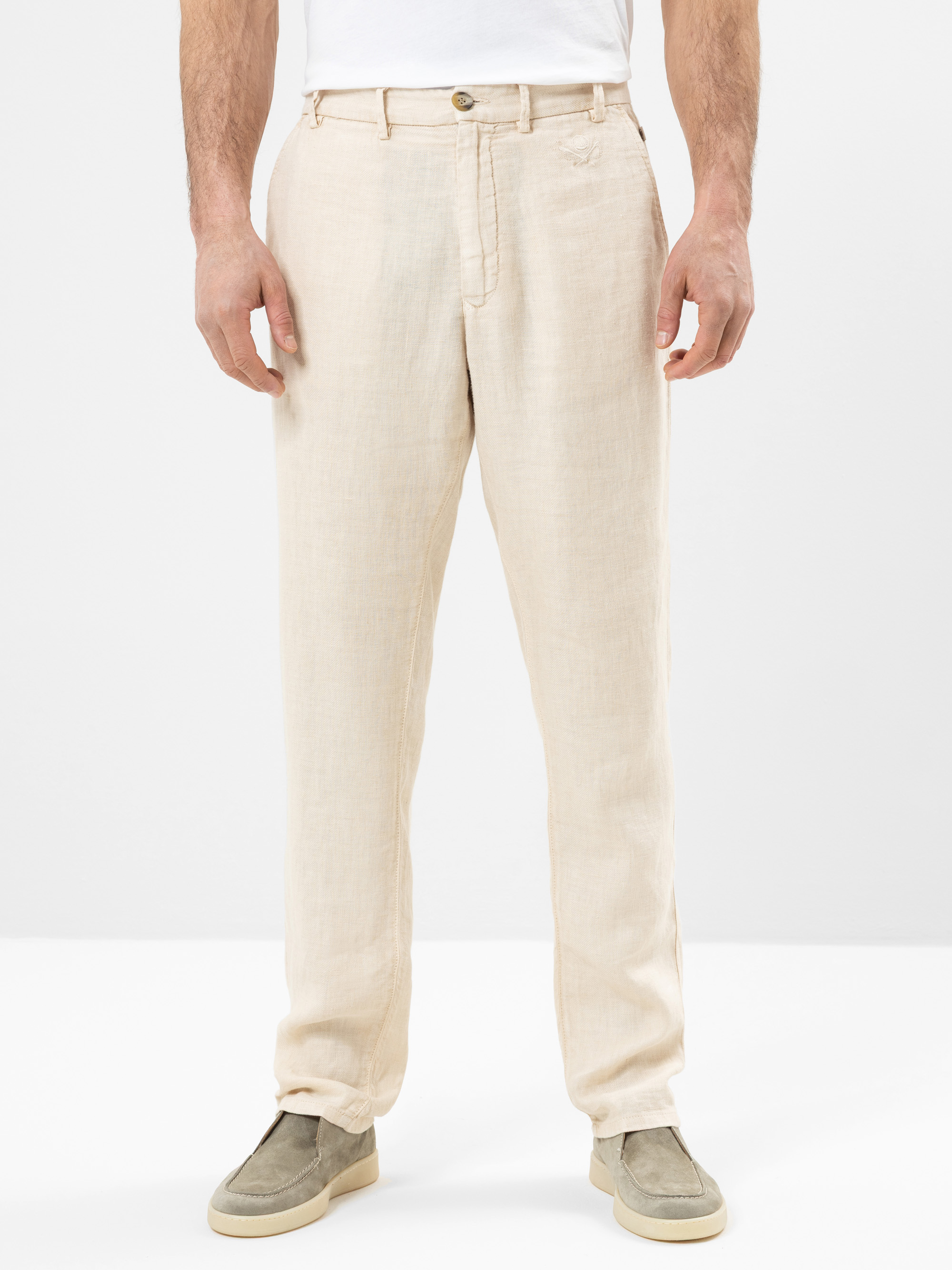 Eine Person, die eine hellbeige Leinenhose, ein weißes Hemd und hellgraue Slipper trägt, steht vor einem schlichten weißen Hintergrund. Das Gesicht der Person ist auf dem Bild nicht zu sehen.