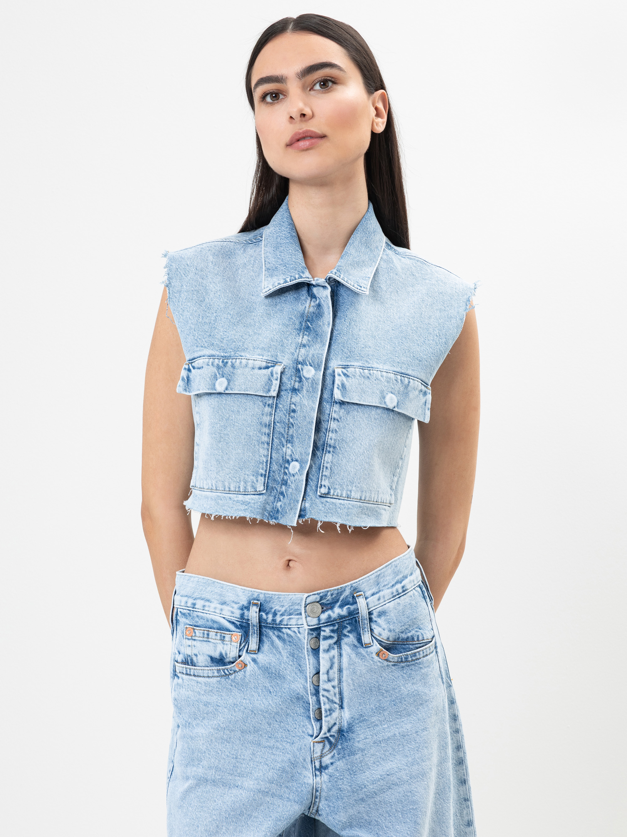 Eine Frau mit langen dunklen Haaren trägt eine ärmellose, hellblaue Jeansweste und hochgekrempelte Jeans, die vor einem schlichten weißen Hintergrund stehen.