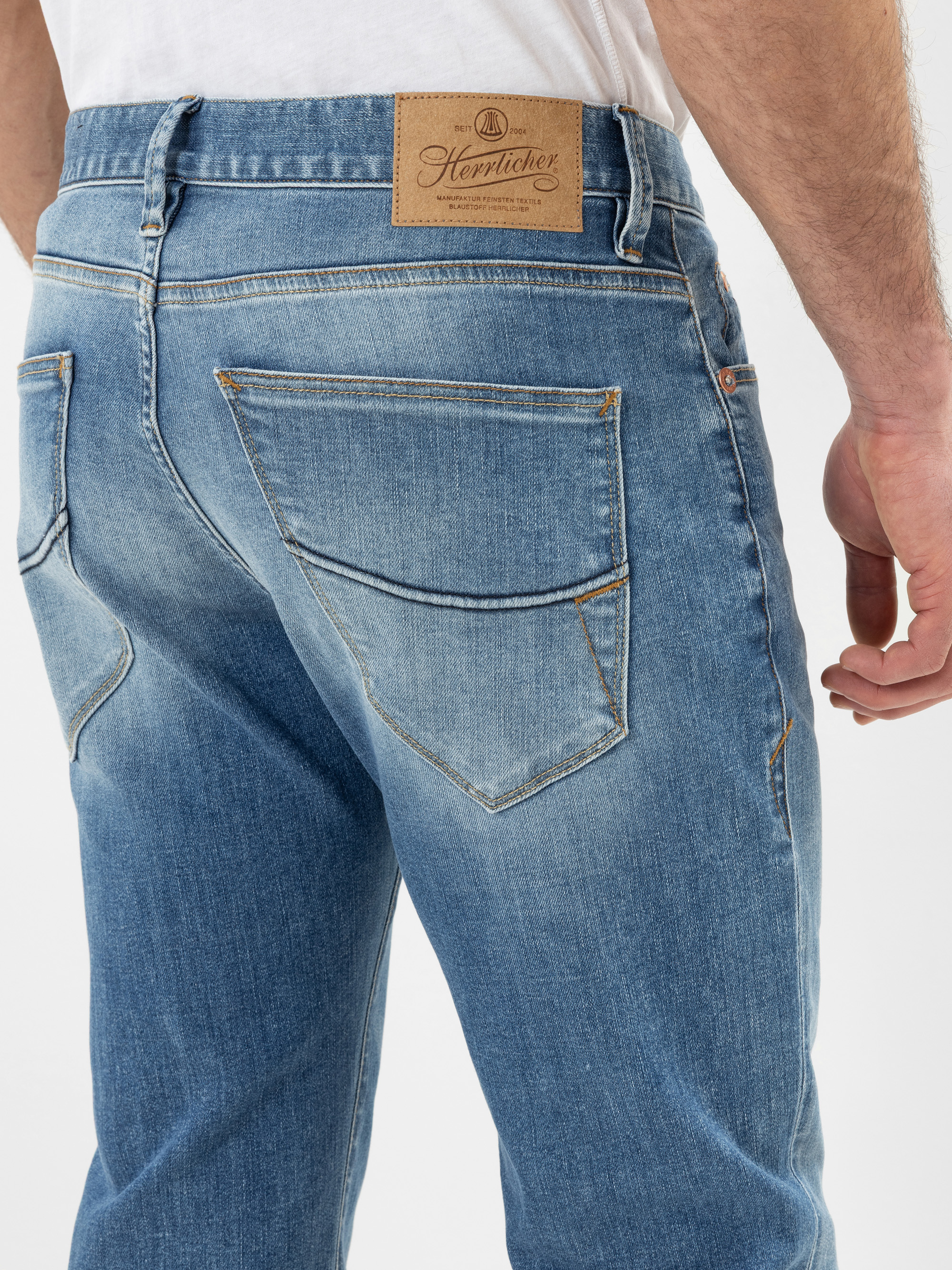 Nahaufnahme einer Person, die eine verblichene blaue Jeans und ein weißes Hemd trägt. Man sieht die Gesäßtaschen und einen braunen Lederaufnäher mit der Aufschrift Herntocher am Hosenbund.