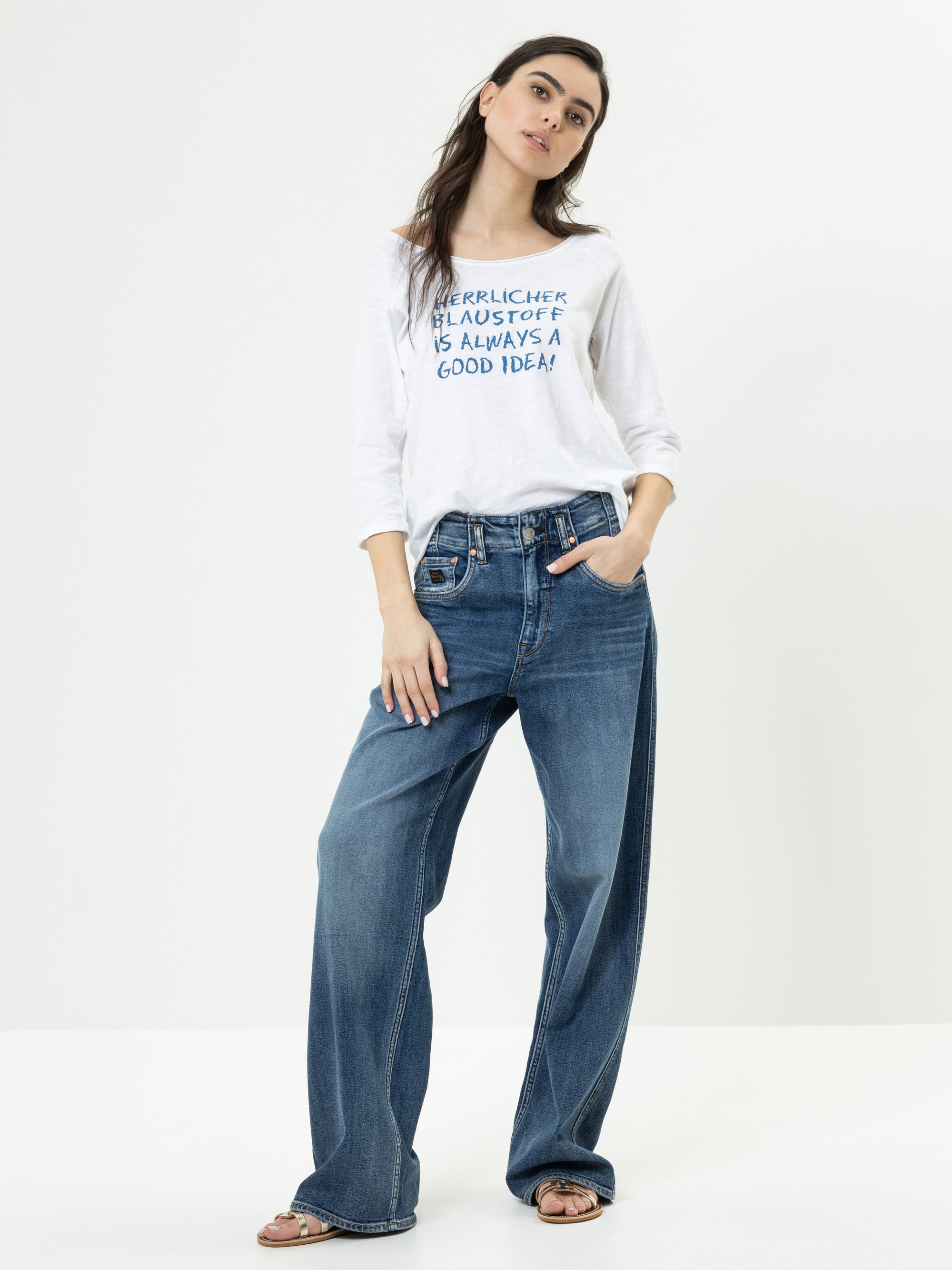 Eine Frau steht vor einem weißen Hintergrund und trägt ein weißes, langärmeliges Hemd mit blauem Text, blaue, weit geschnittene Jeans und Sandalen. Sie posiert mit einer Hand in der Tasche und einem entspannten Gesichtsausdruck.