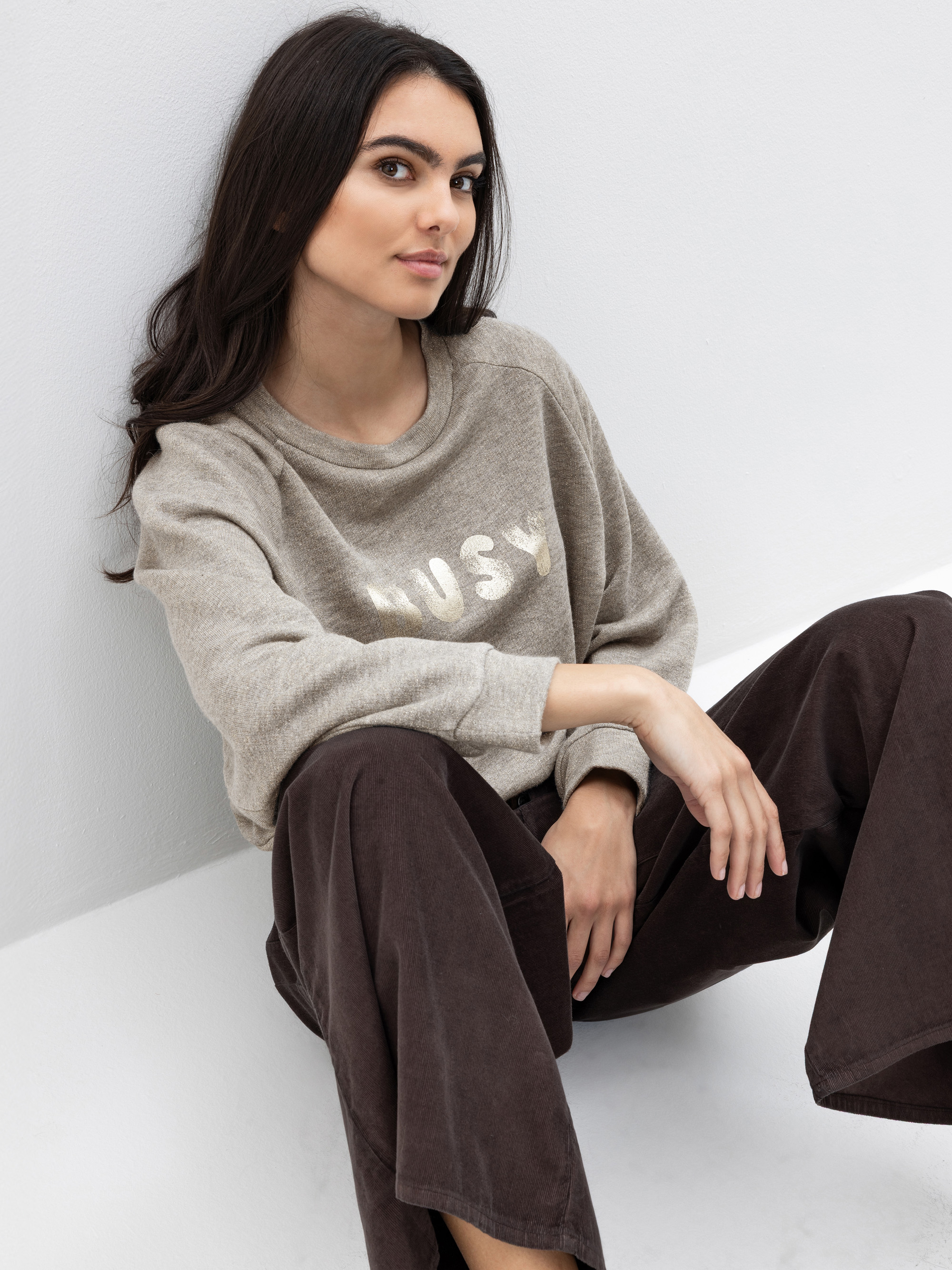 Eine Frau mit langen dunklen Haaren sitzt auf dem Boden vor einer weißen Wand. Sie trägt ein hellgraues Sweatshirt mit der Aufschrift BUSY und eine weite dunkelbraune Hose. Sie blickt mit einem leichten Lächeln in die Kamera.