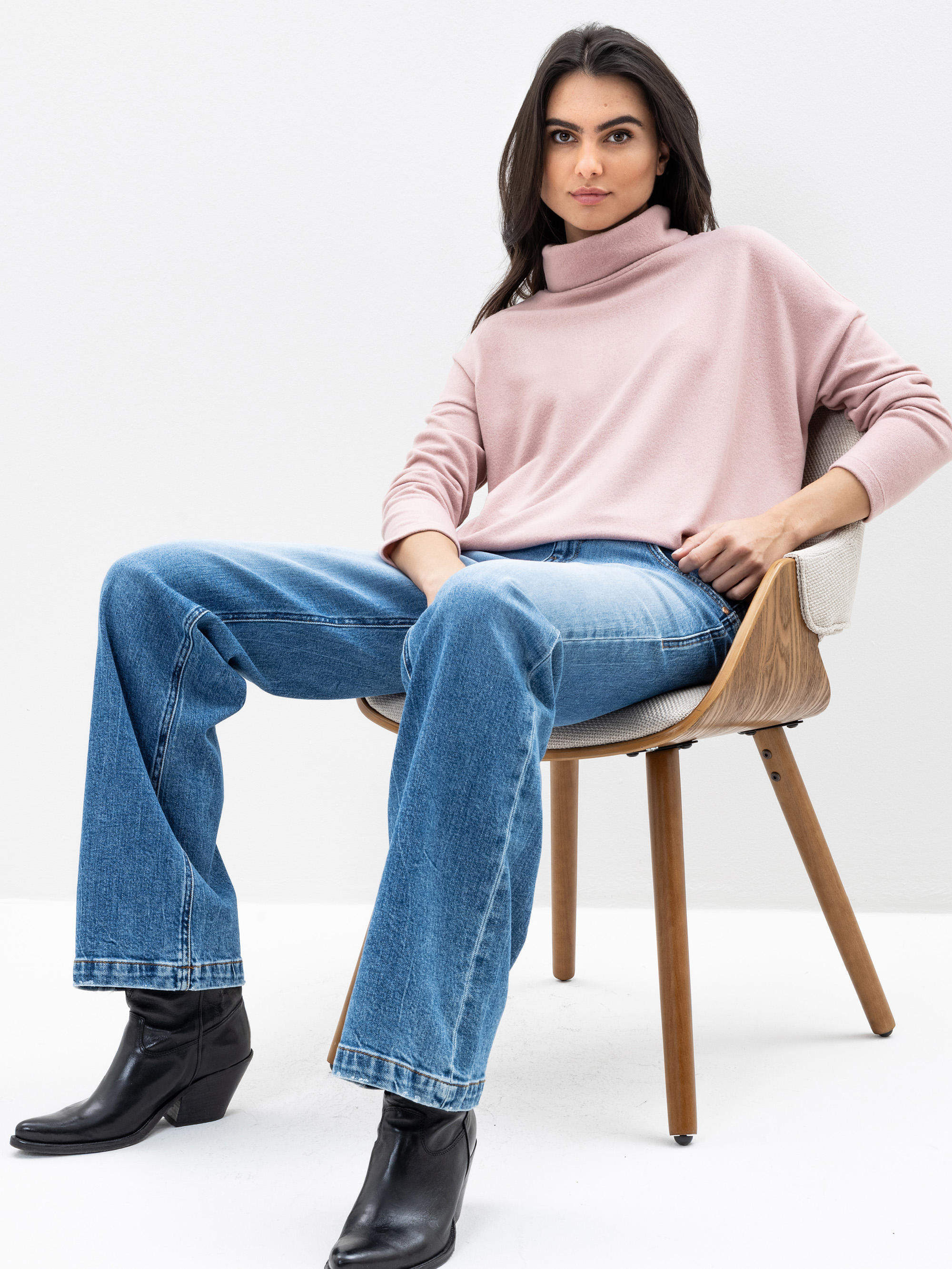 Eine Frau sitzt auf einem modernen Holzstuhl, trägt einen hellrosa Rollkragenpullover, blaue Jeans und schwarze Stiefeletten. Sie blickt selbstbewusst in die Kamera vor einem schlichten weißen Hintergrund.