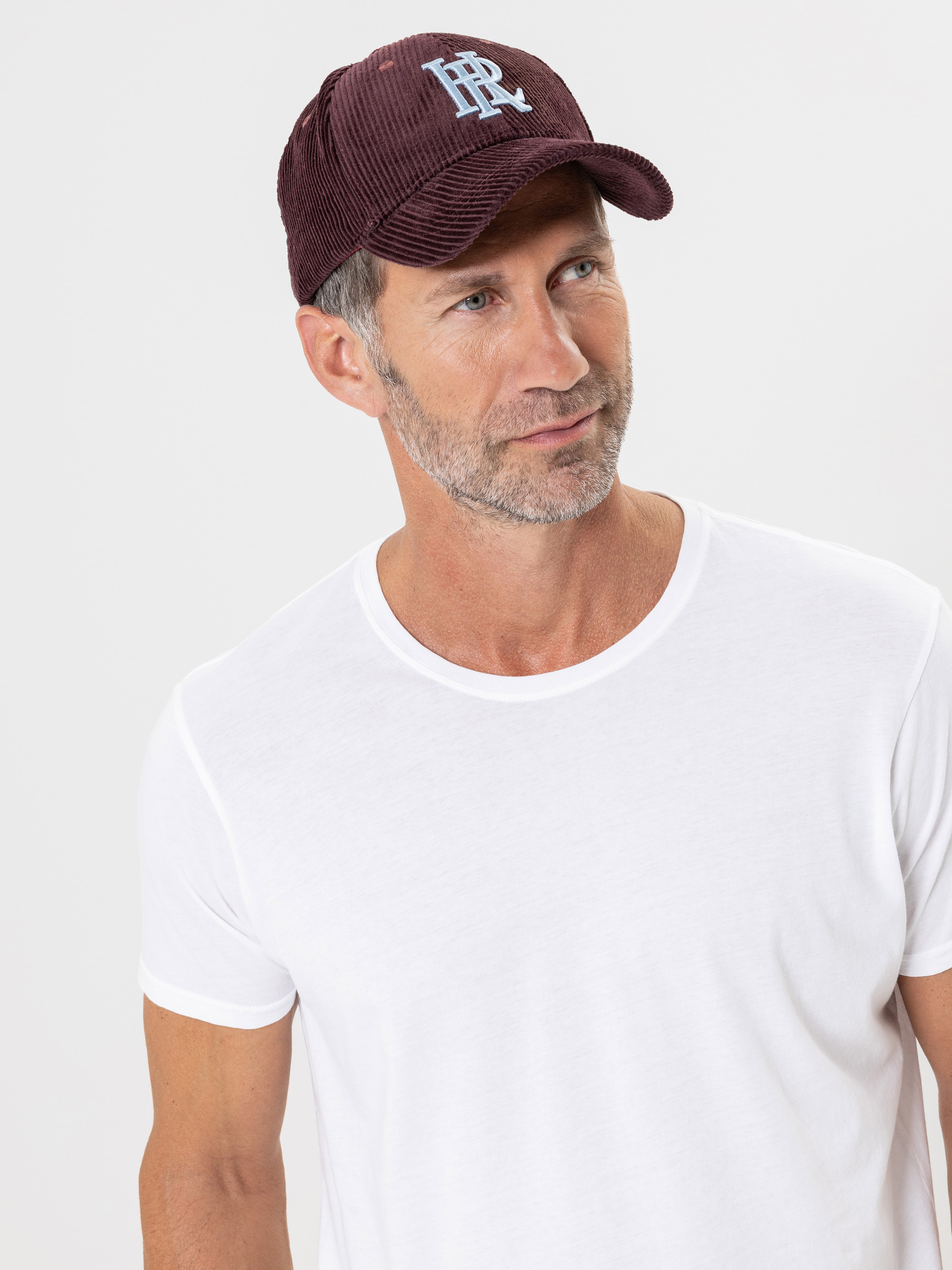 Ein Mann mit heller Haut, kurzen braunen Haaren und einem Bart trägt ein weißes T-Shirt und eine kastanienbraune Baseballmütze mit hellblauer Stickerei und steht vor einem schlichten weißen Hintergrund.