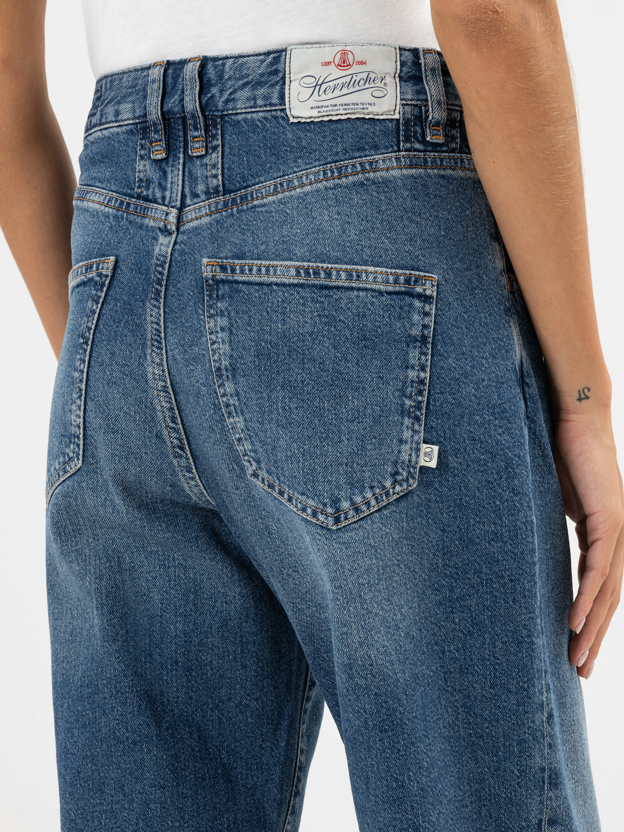 Nahaufnahme einer Person, die eine blaue Jeans mit hoher Taille und einem weißen Aufnäher mit der Aufschrift "Hereficker" am Bund trägt. Die Gesäßtasche und ein Teil der Hand der Person mit einer kleinen Tätowierung am Handgelenk sind sichtbar.