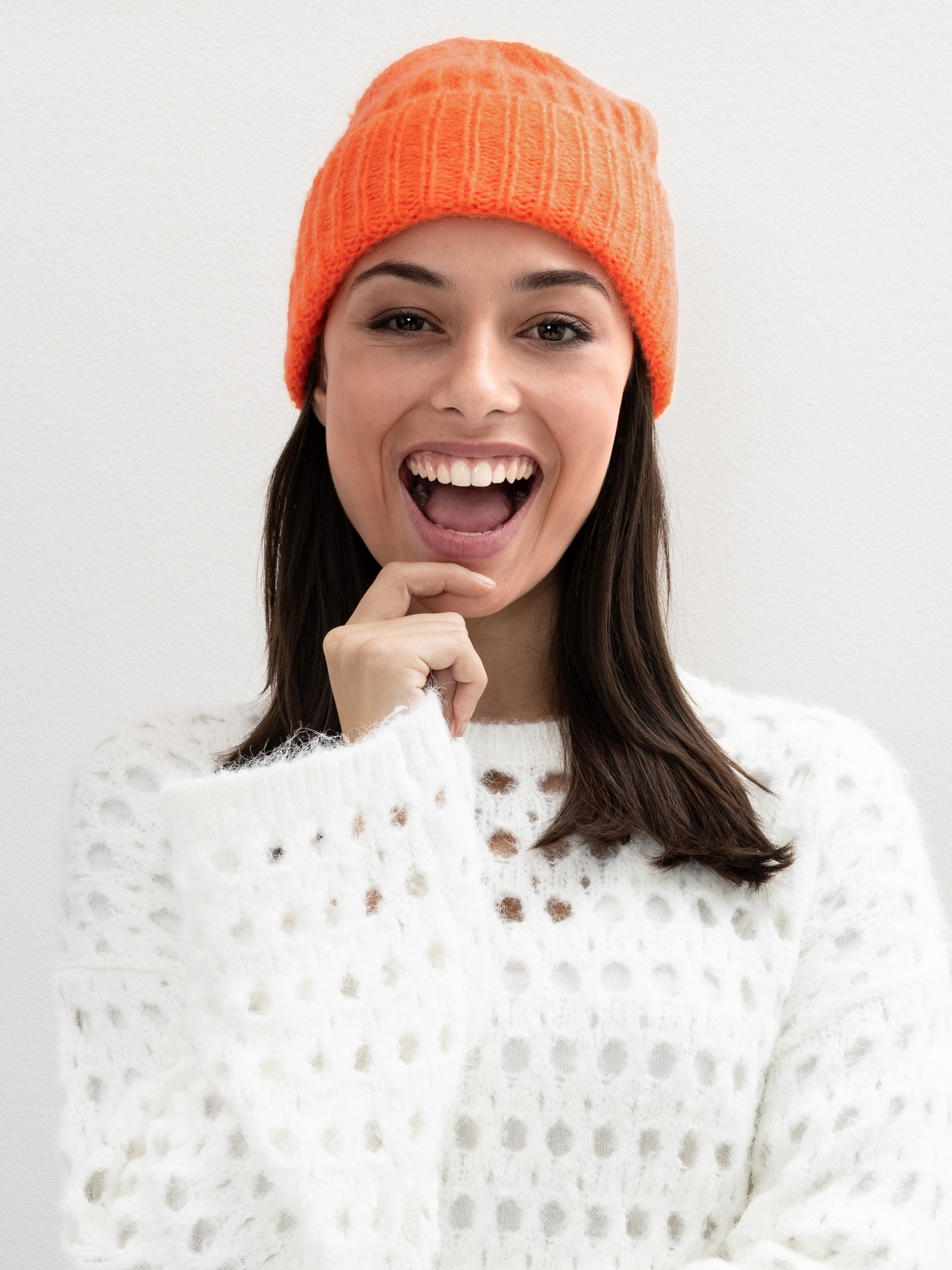 Eine Frau, die eine orangefarbene Strickmütze und einen weißen strukturierten Pullover trägt, lächelt mit offenem Mund und der Hand am Kinn vor einem schlichten hellen Hintergrund.