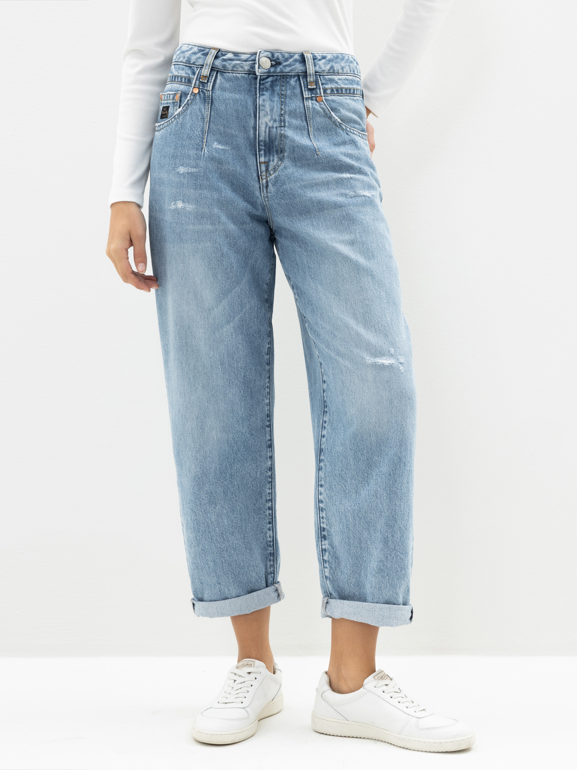 Eine Person, die eine hellblaue, hochtaillierte, locker sitzende Jeans mit aufgerollten Bündchen, ein weißes langärmeliges Oberteil und weiße Turnschuhe trägt, steht vor einem schlichten weißen Hintergrund.