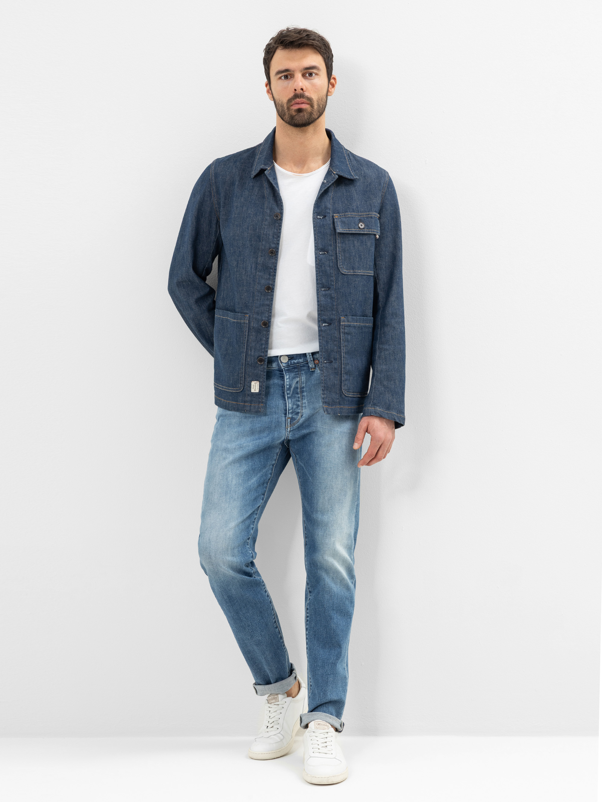 Ein Mann mit kurzen dunklen Haaren und Bart, der eine blaue Jeansjacke über einem weißen T-Shirt, hellblaue Jeans und weiße Turnschuhe trägt, steht mit übereinander geschlagenen Beinen vor einer schlichten weißen Wand.