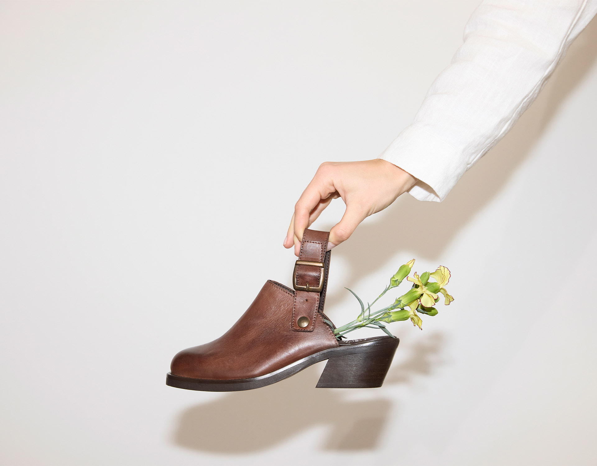 Eine Hand in einem weißen Ärmel hält einen braunen Schuh mit Absatz an seinem Riemen; gelbe Blumen mit grünen Stielen sind im Inneren des Schuhs vor einem schlichten hellen Hintergrund angeordnet.