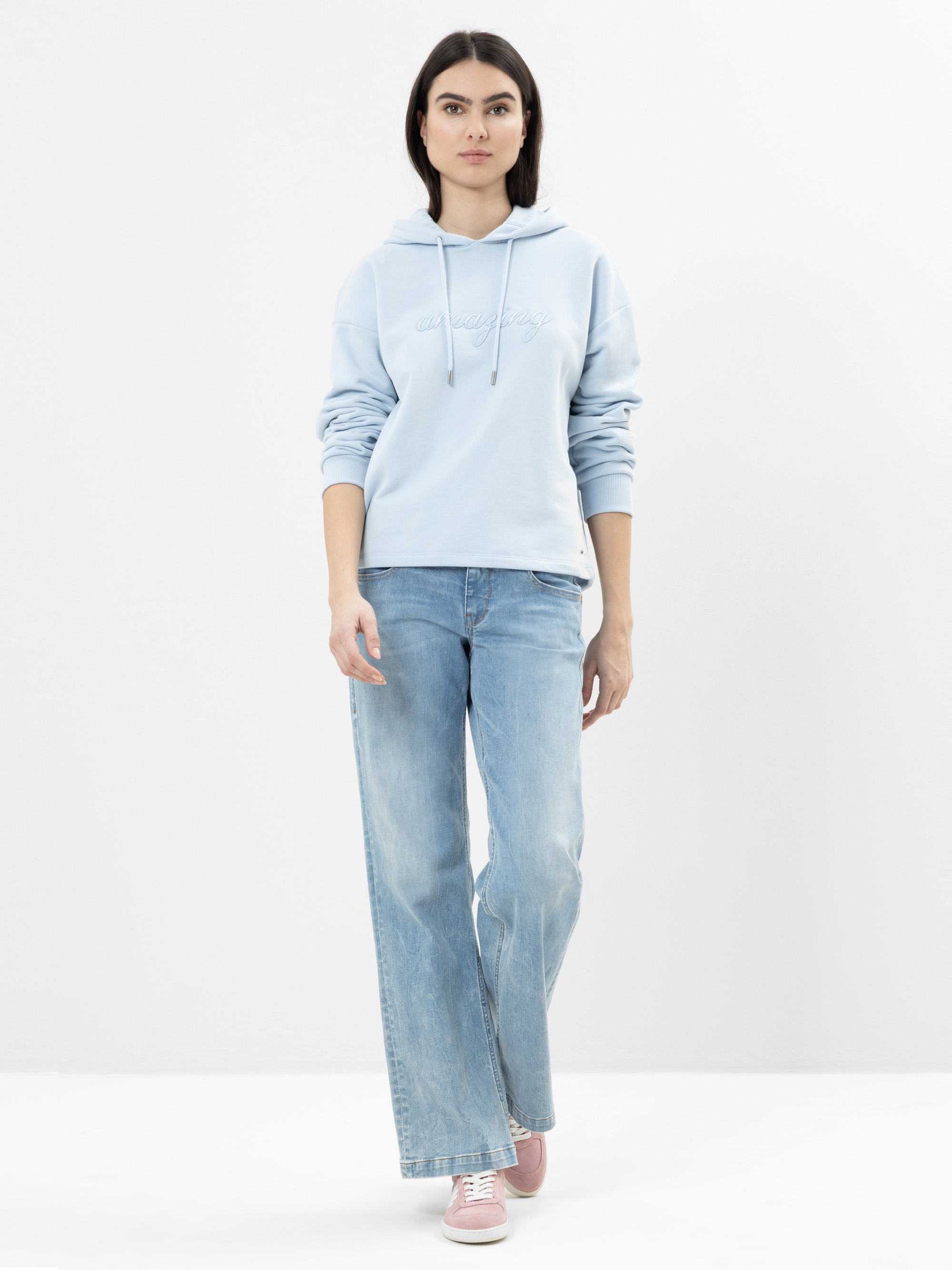 Eine Person mit langen dunklen Haaren trägt einen hellblauen Kapuzenpulli, hellblaue Jeans mit weitem Bein und rosa Turnschuhe und steht vor einem schlichten weißen Hintergrund.