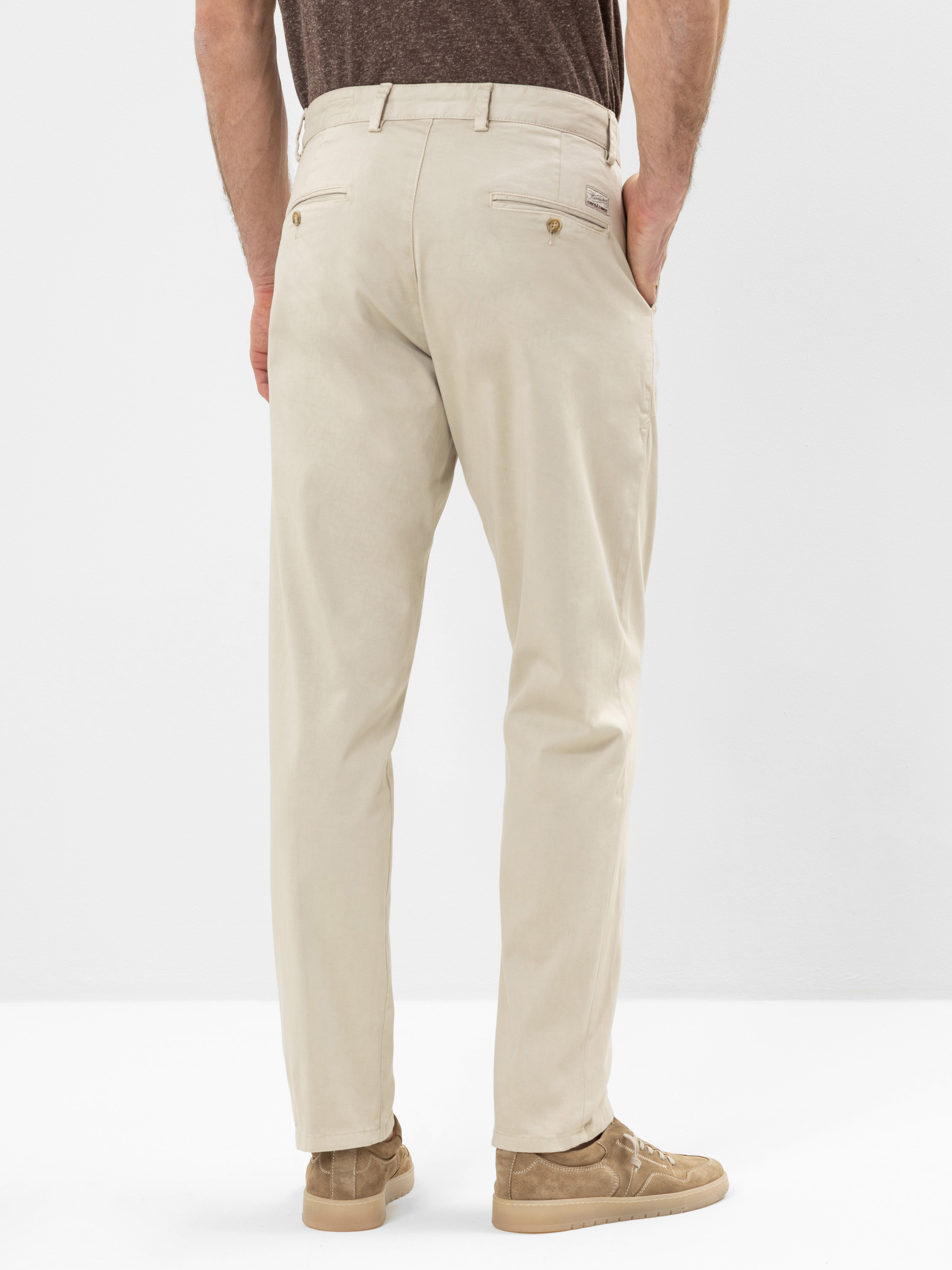 Ein Mann steht mit dem Gesicht zur Wand und trägt eine hellbeige Chinohose, ein braunes T-Shirt und hellbraune Wildlederschuhe. Der Hintergrund ist schlicht und weiß.