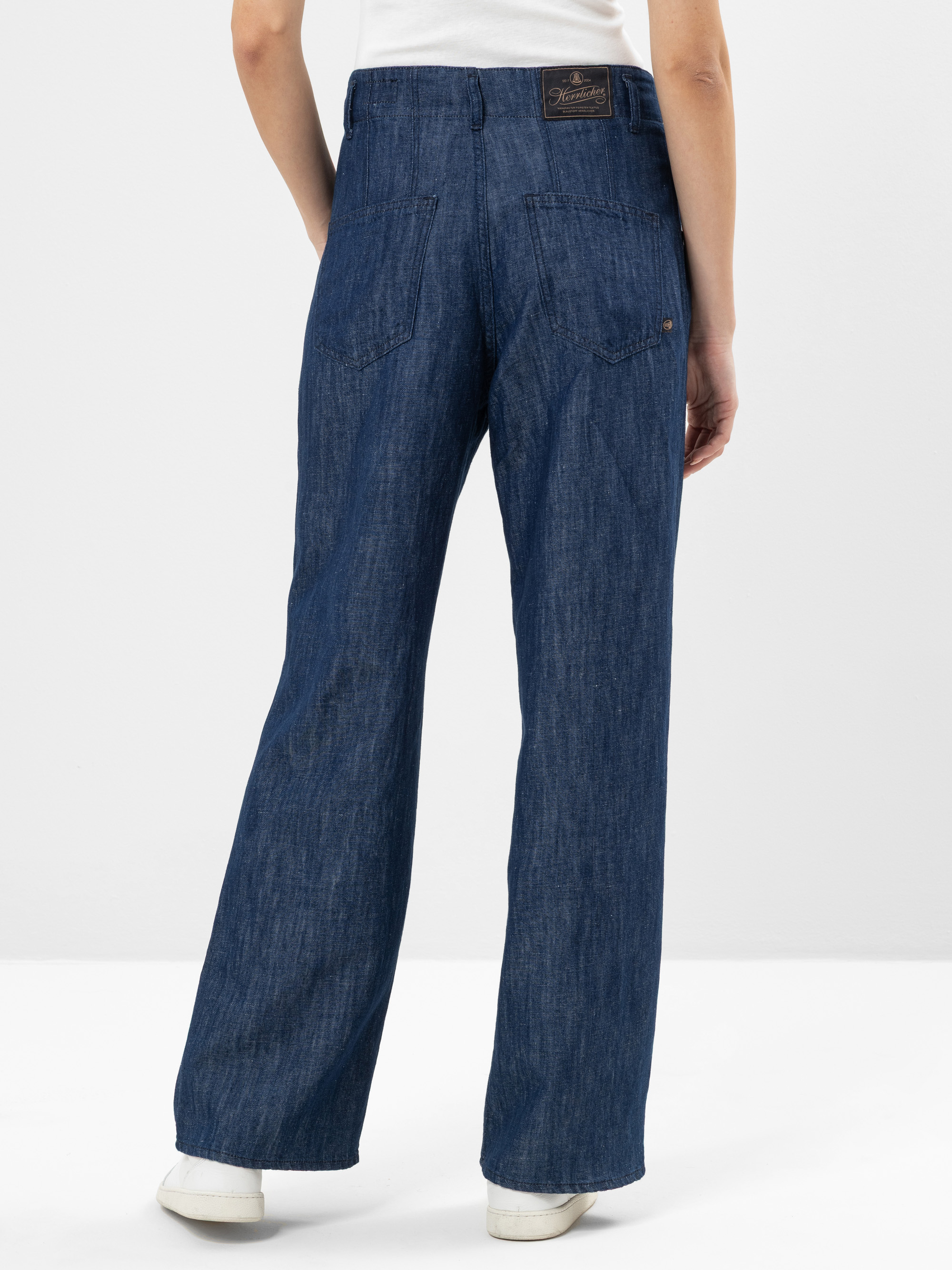 Stehende Person vor weißem Hintergrund, von hinten gesehen, mit weit geschnittenen blauen Jeans, weißen Turnschuhen und einem weißen ärmellosen Oberteil. Die Jeans hat zwei Gesäßtaschen und ein dunkelbraunes Etikett am Bund.