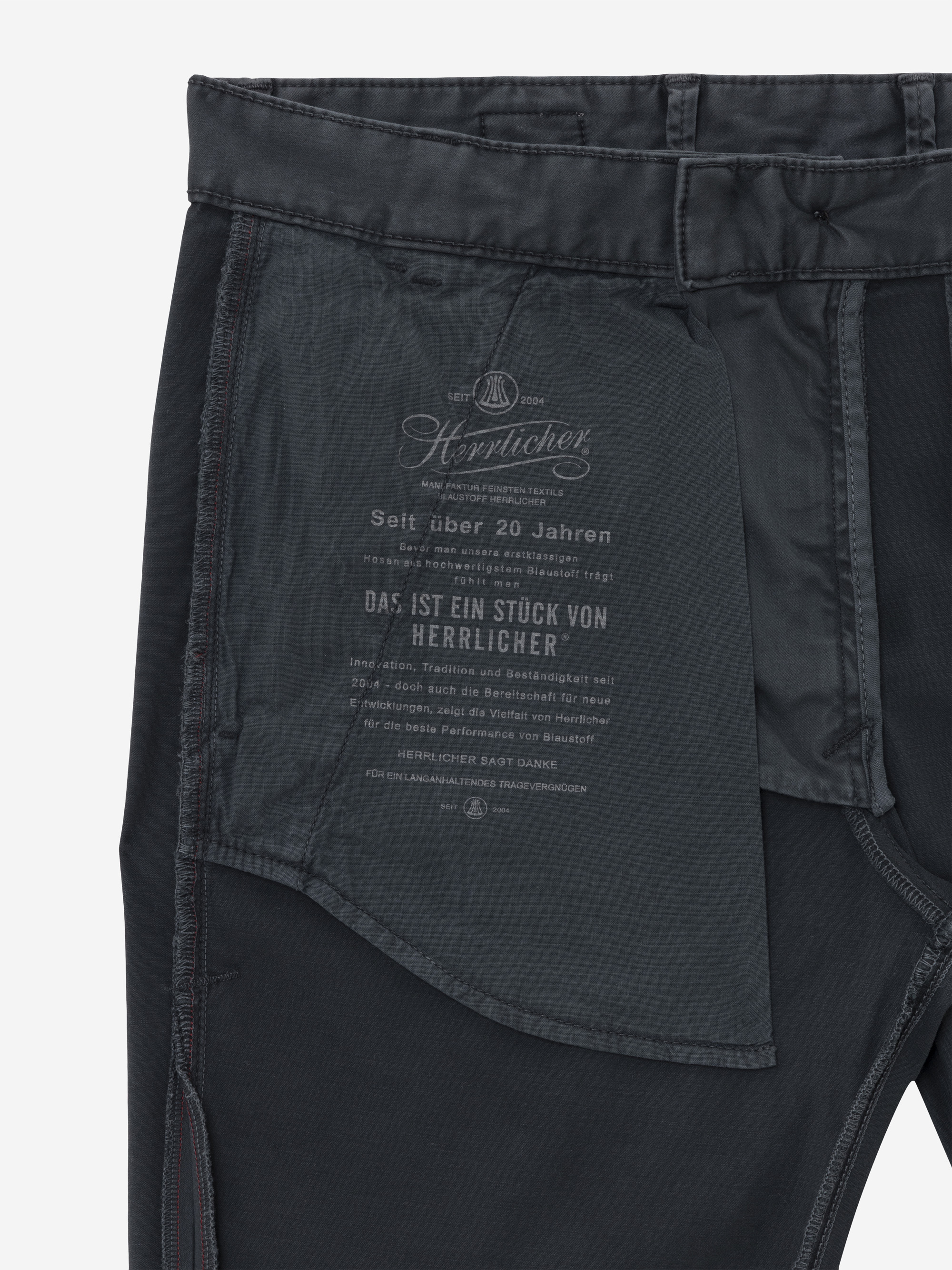 Nahaufnahme der Innenseite einer dunklen Hose mit Markentext und einem auf das Taschenfutter aufgedruckten Logo in deutscher Sprache, das auf das Erbe und die Handwerkskunst der Bekleidungsmarke Herrlicher hinweist.