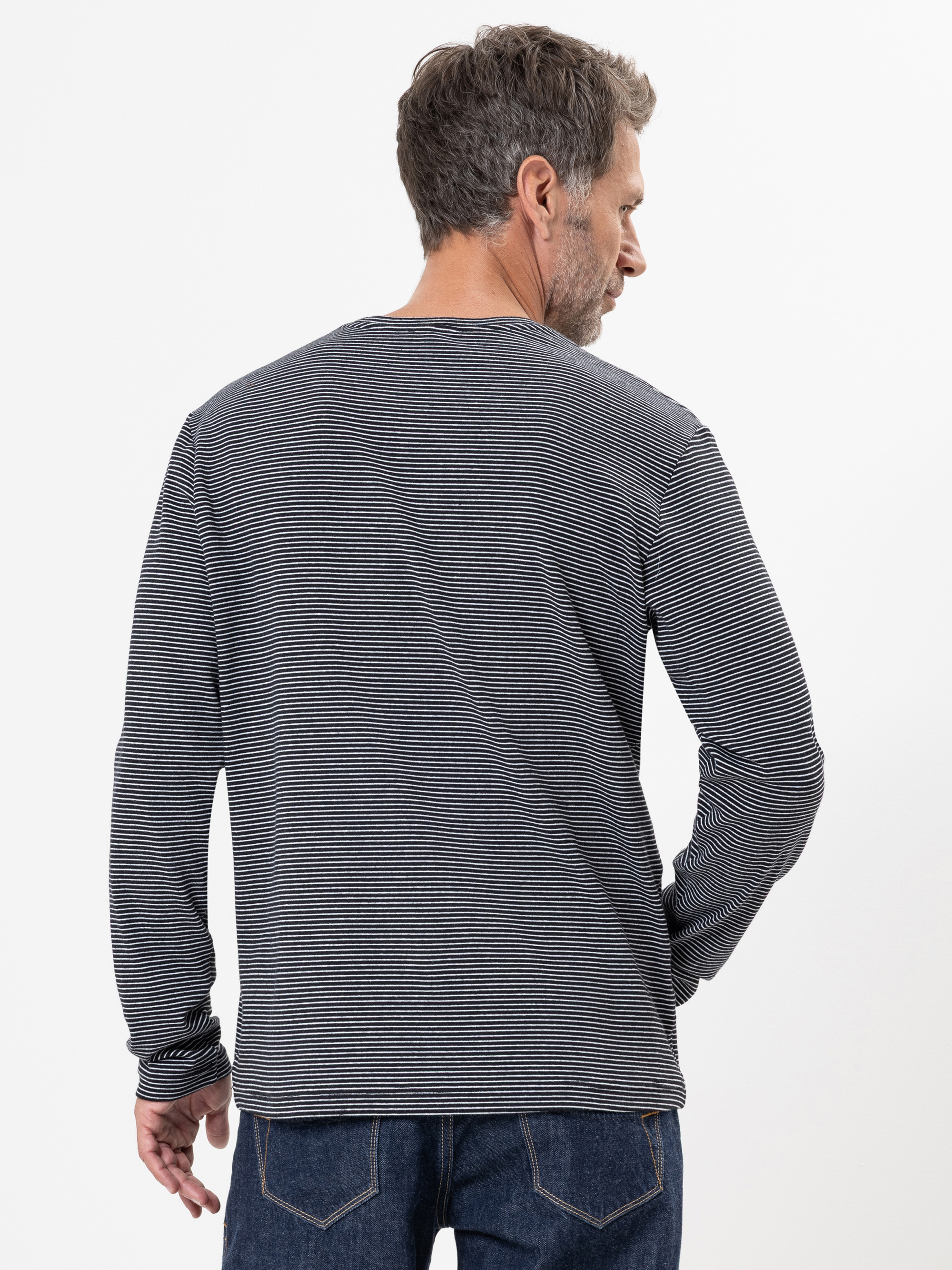 Ein Mann mit kurzen Haaren steht mit dem Rücken zur Kamera und trägt ein langärmeliges, schwarz-weiß gestreiftes Hemd und blaue Jeans vor einem schlichten weißen Hintergrund.