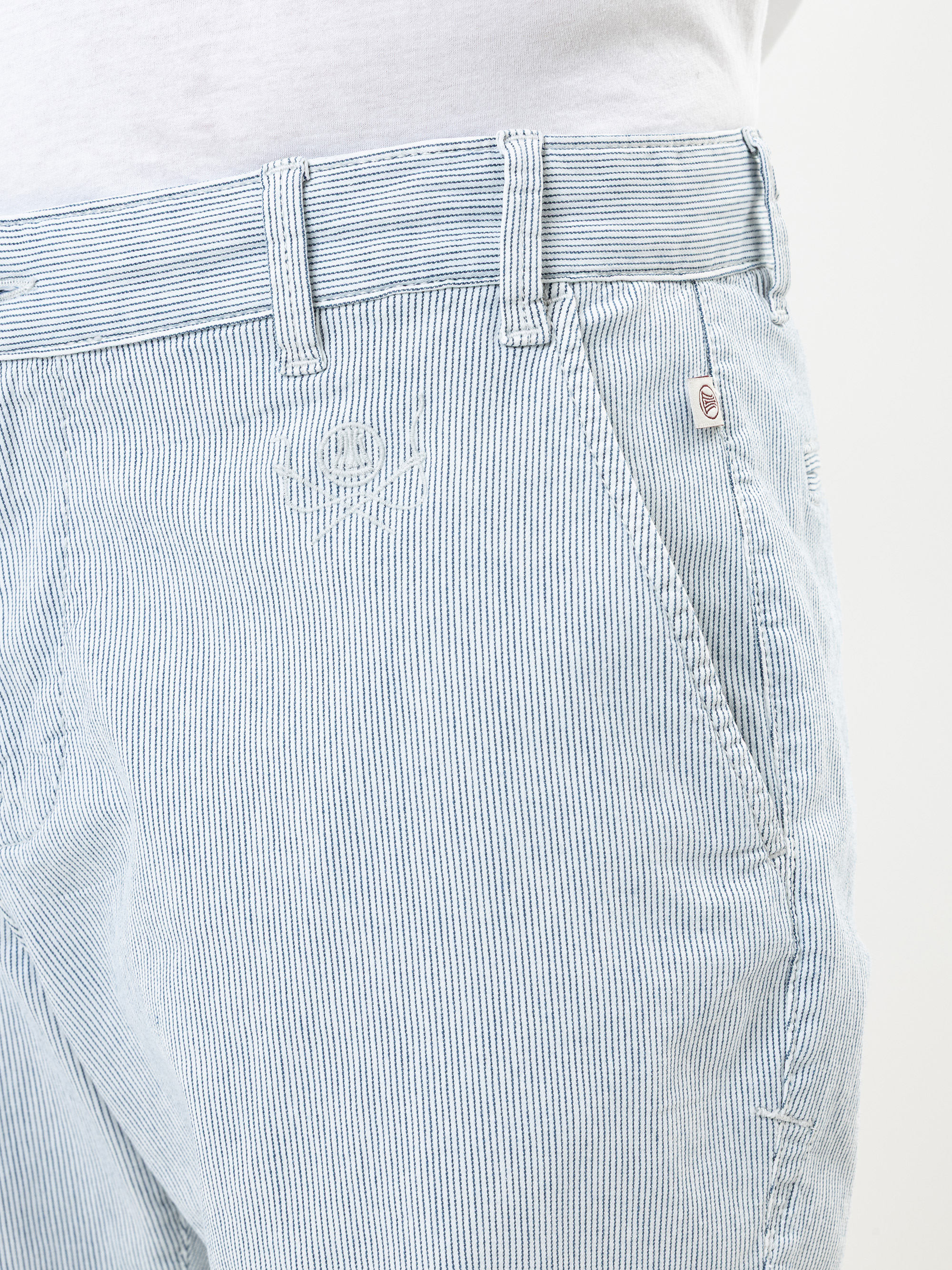 Nahaufnahme einer Person, die eine hellblau-weiß gestreifte Hose und ein weißes Hemd mit Stecktuch trägt. Die Hose hat Gürtelschlaufen und eine Vordertasche mit einer kleinen Metallknopfleiste.
