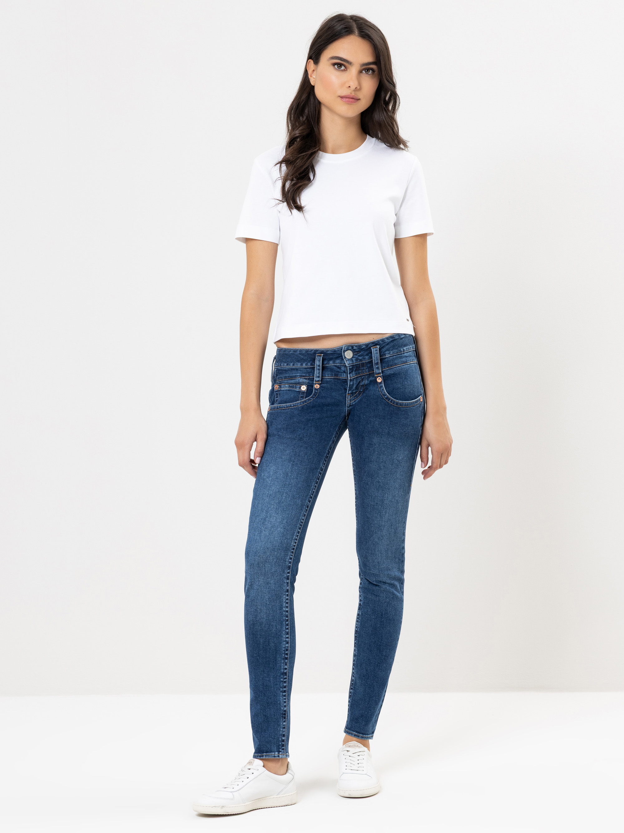 Eine Frau steht vor einem schlichten weißen Hintergrund und trägt ein weißes, kurzärmeliges T-Shirt, blaue Skinny Jeans und weiße Turnschuhe. Sie hat langes, gewelltes dunkles Haar und einen neutralen Gesichtsausdruck.