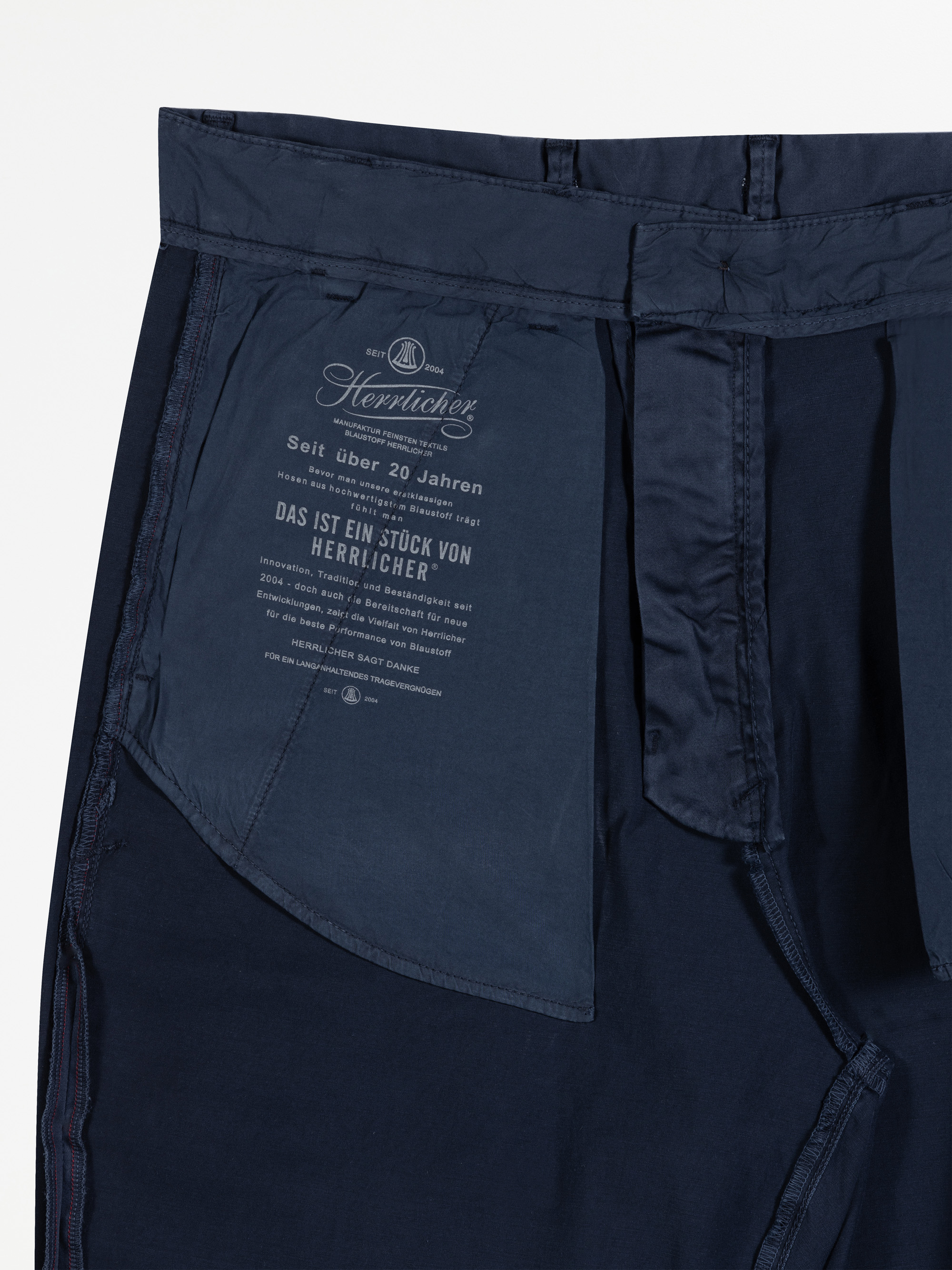 Nahaufnahme der Innenseite einer marineblauen Hose, die die Innentasche mit aufgedrucktem deutschen Text zeigt, einschließlich Markenzeichen und Pflegeinformationen der Bekleidungsfirma Herrlicher.