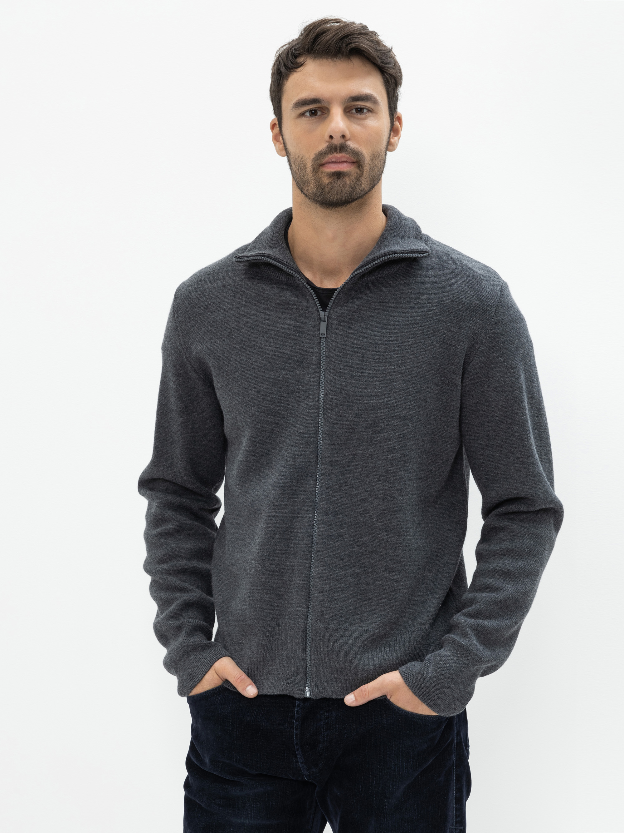 Ein Mann mit kurzen dunklen Haaren und einem Bart trägt einen dunkelgrauen Pullover mit Reißverschluss und eine dunkle Hose. Er steht vor einem schlichten weißen Hintergrund, hat die Hände in den Taschen und schaut in die Kamera.