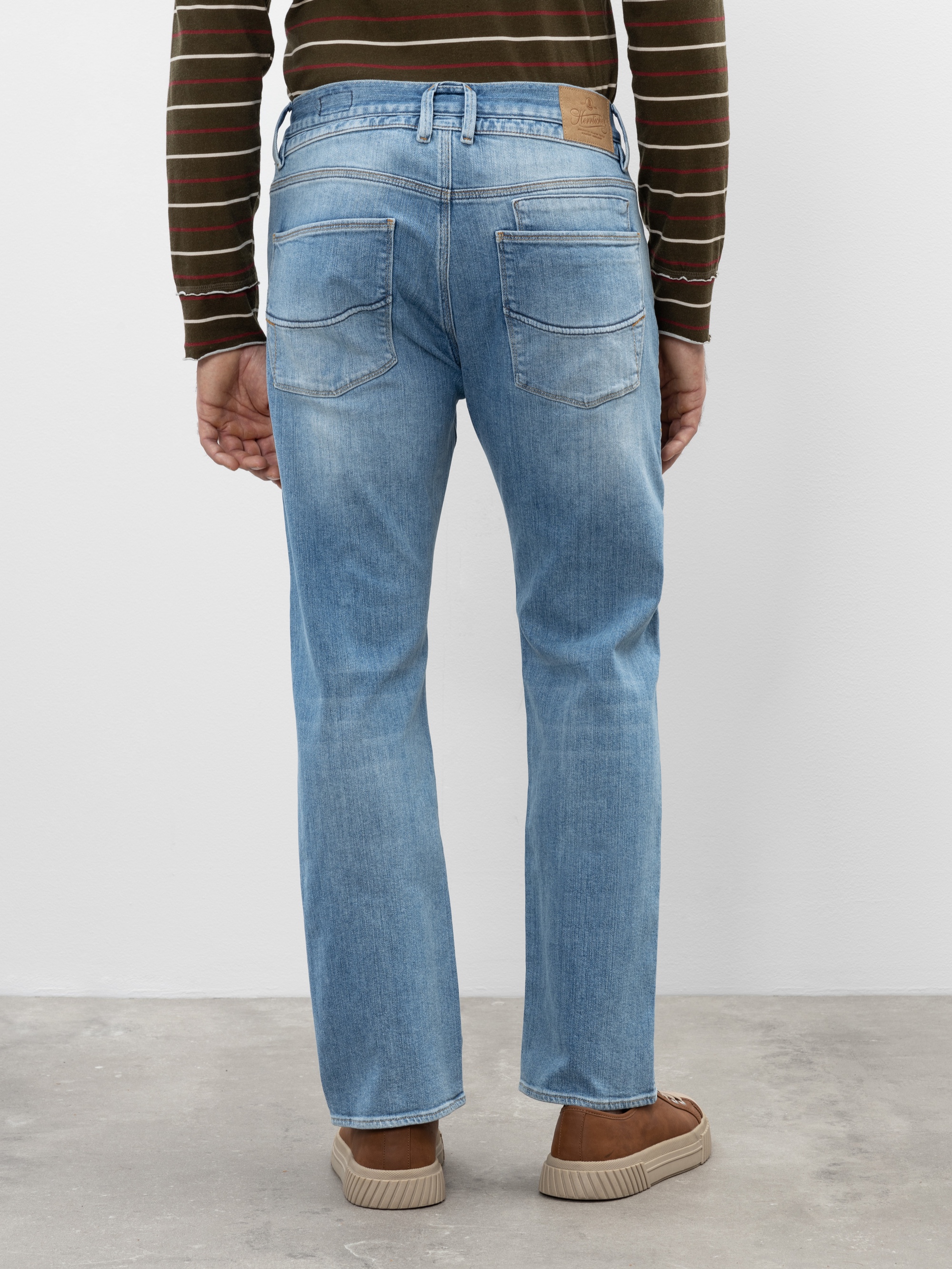 Eine Person, die hellblaue Jeans, ein gestreiftes langärmeliges Hemd und braune Turnschuhe trägt, steht mit dem Gesicht zur Kamera vor einer hellen Wand und einem Betonboden.