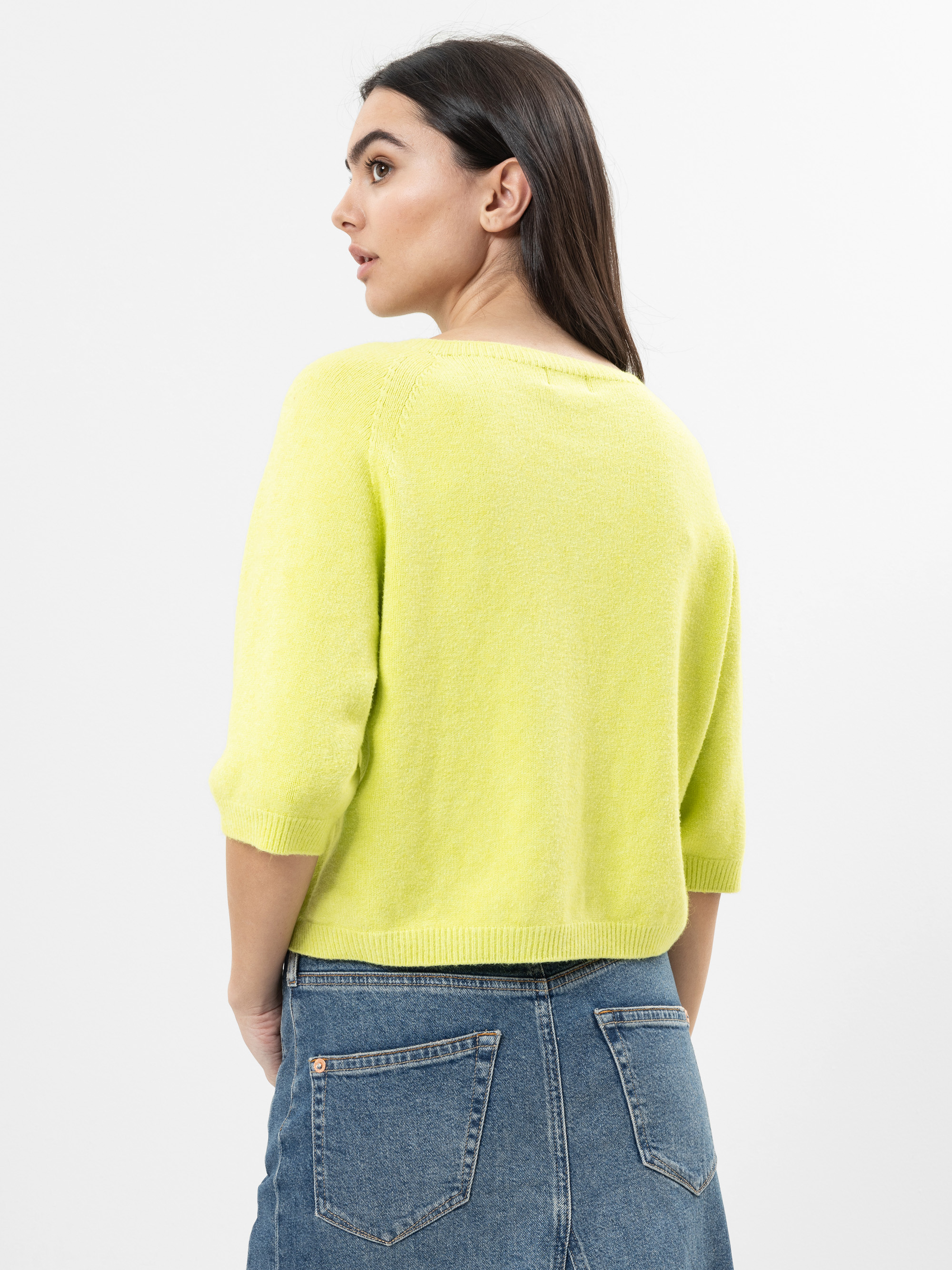 Eine Frau mit langen dunklen Haaren trägt einen leuchtend gelben Pullover mit Dreiviertelärmeln und einen blauen Jeansrock, von hinten und leicht zur Seite gedreht, vor einem schlichten weißen Hintergrund.