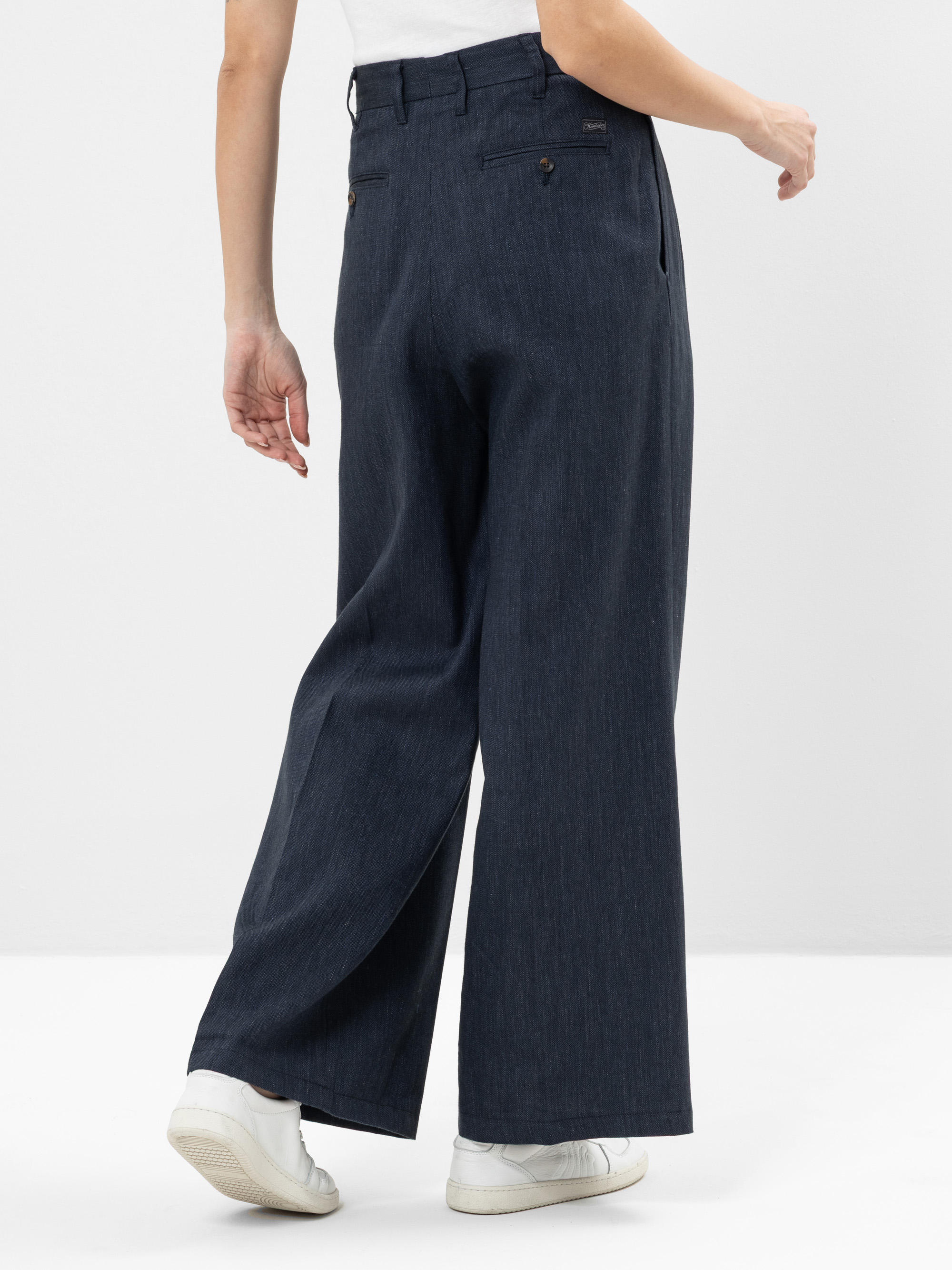 Person, die eine dunkelblaue Hose mit weitem Bein, ein weißes Oberteil und weiße Turnschuhe trägt, fotografiert von hinten vor einem schlichten weißen Hintergrund.