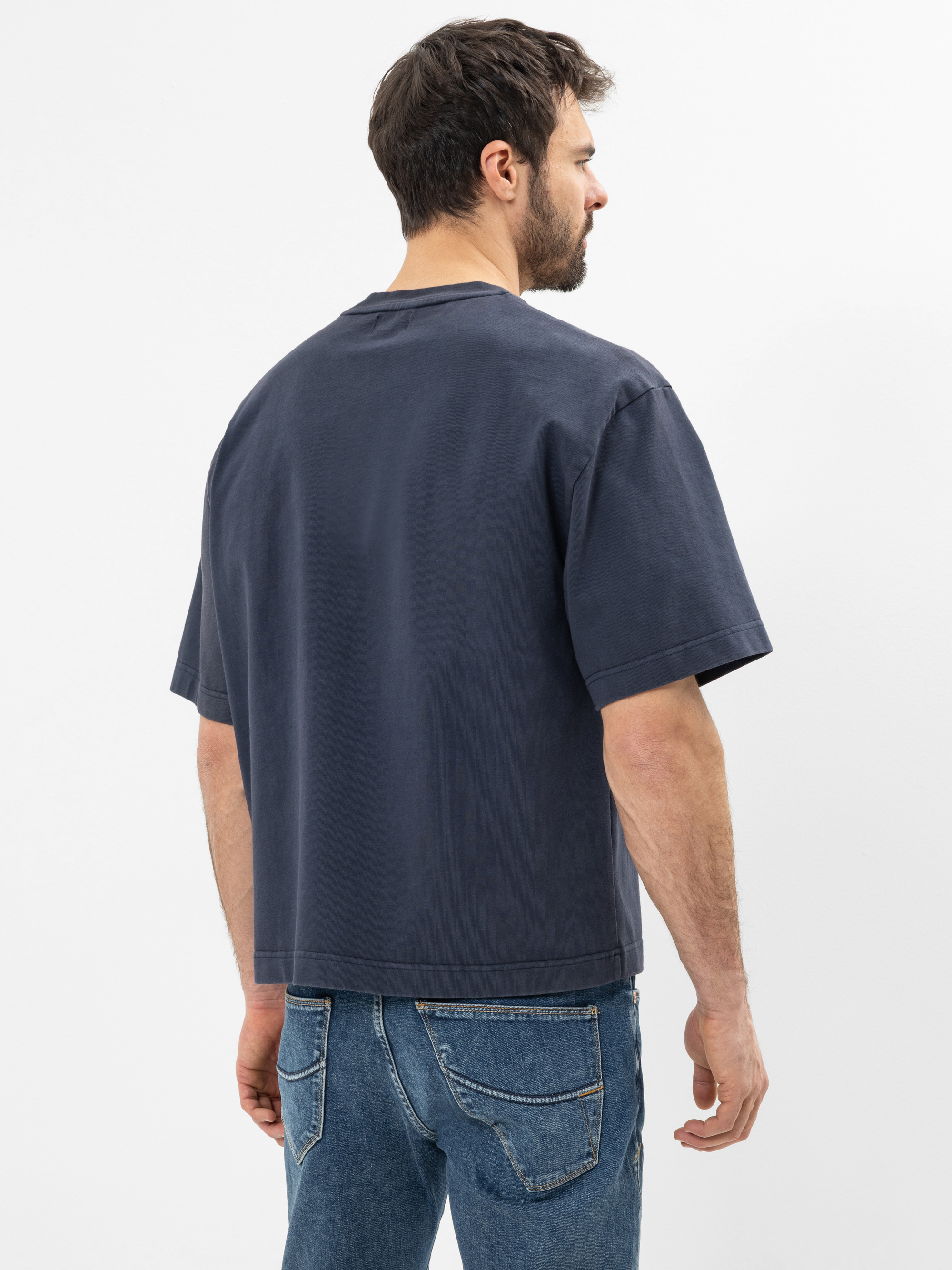 Ein Mann mit kurzen dunklen Haaren und einem Bart steht mit dem Rücken zur Kamera. Er trägt ein dunkelblaues T-Shirt und eine blaue Jeans vor einem schlichten weißen Hintergrund.