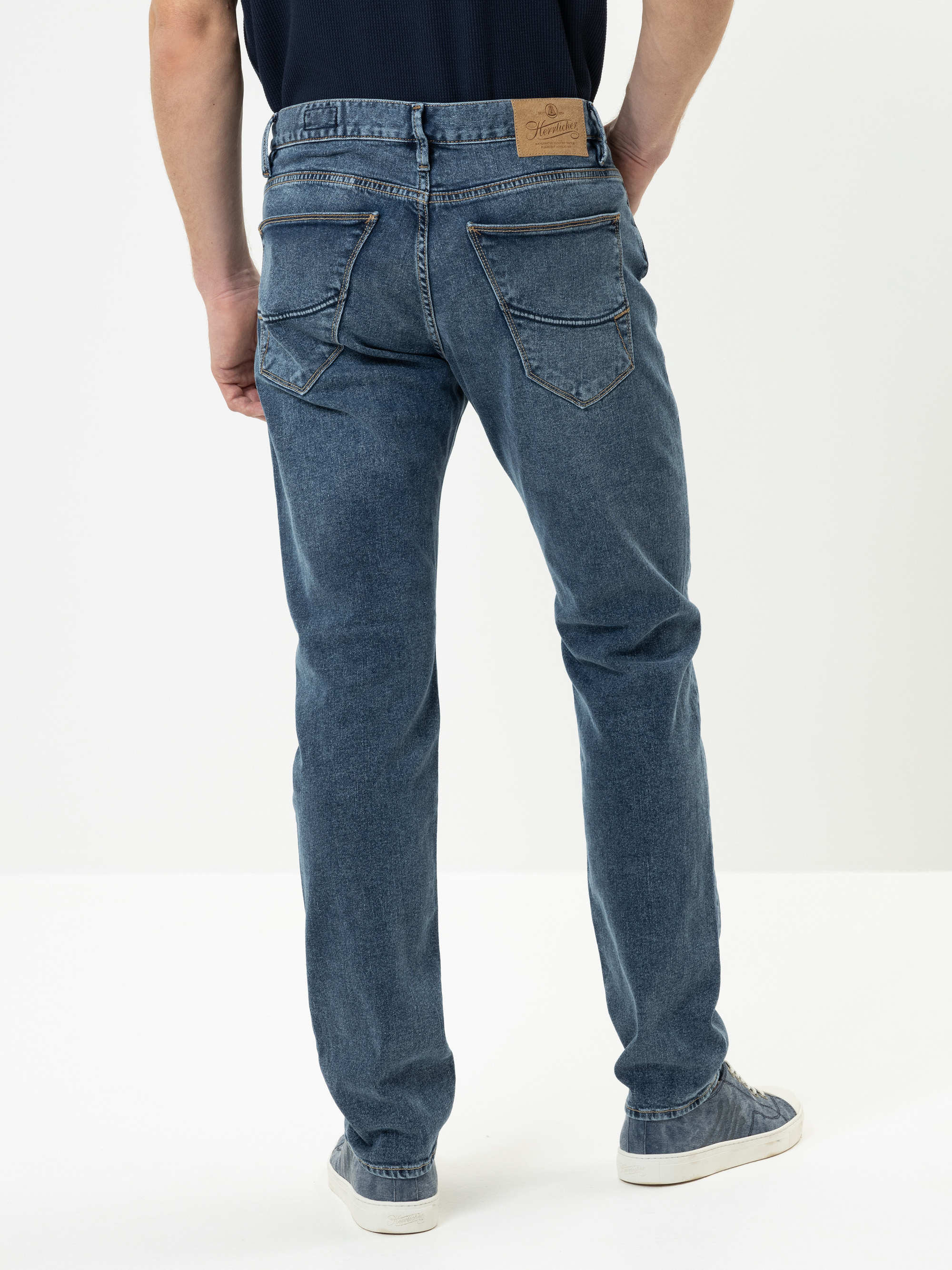 Eine Person, die blaue Jeans und graue Turnschuhe trägt, steht mit dem Rücken zur Kamera vor einem schlichten weißen Hintergrund. Die Jeans hat sichtbare Gesäßtaschen und einen braunen Lederaufnäher am Bund.