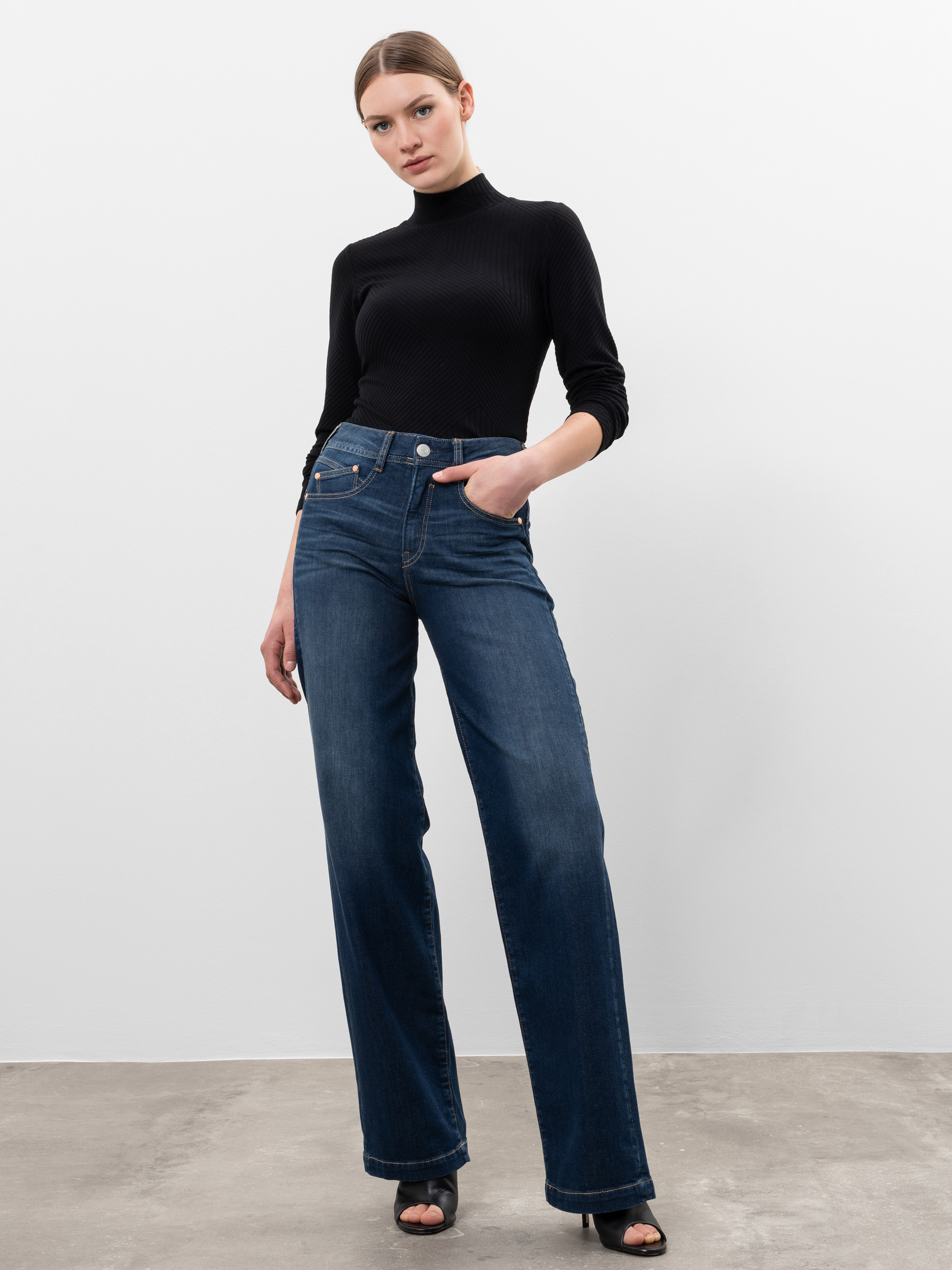 Eine Frau steht vor einer schlichten weißen Wand, trägt ein schwarzes Rollkragenoberteil, eine hoch sitzende Jeans mit weitem Bein und schwarze, offene Absätze, eine Hand in der Tasche und einen neutralen Gesichtsausdruck.