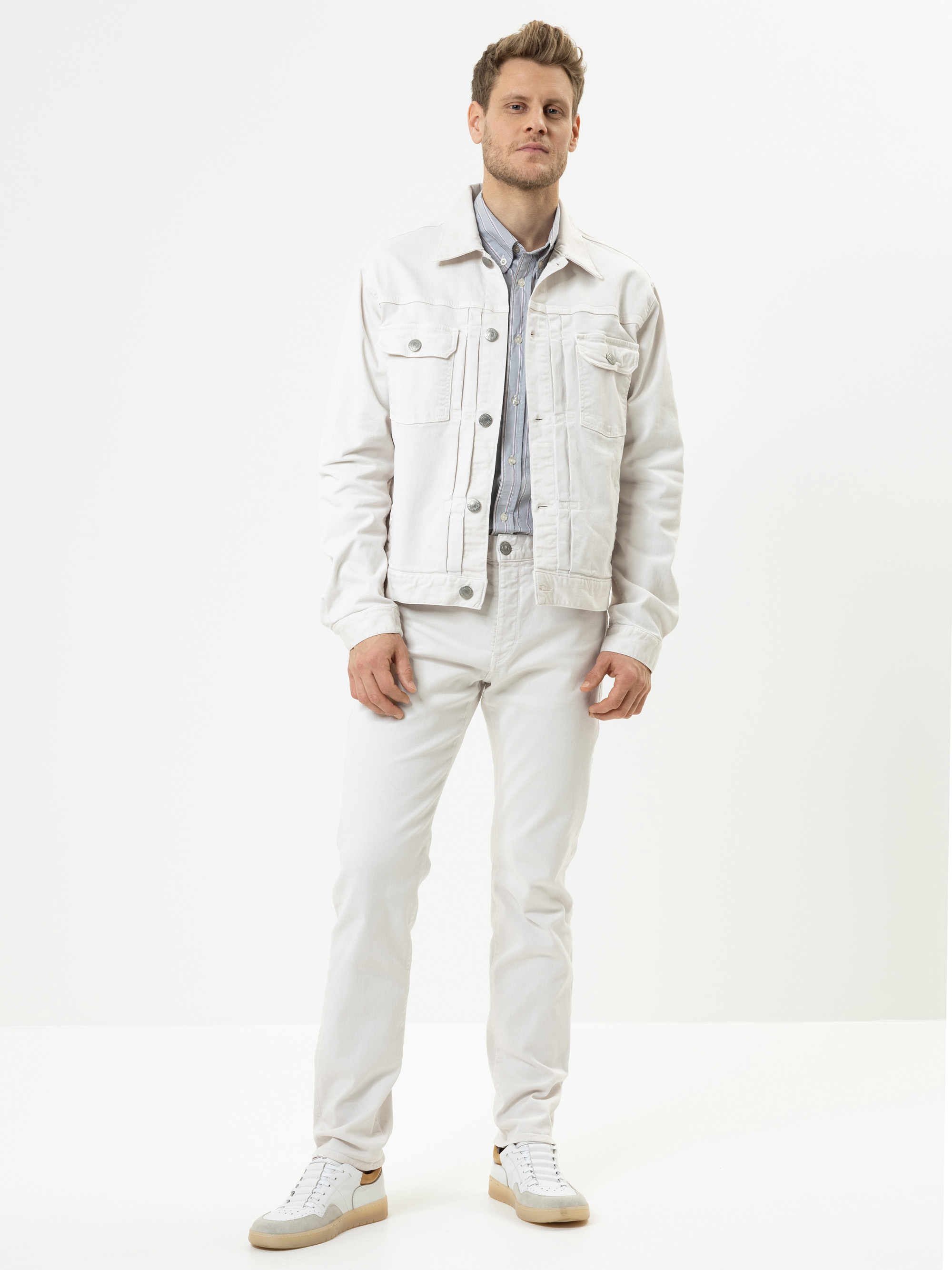 Ein Mann steht vor einem schlichten weißen Hintergrund und trägt eine helle Jeansjacke, passende Jeans, ein graues Hemd und weiße Turnschuhe. Er blickt mit neutraler Miene nach vorne und hat eine Hand in der Tasche.