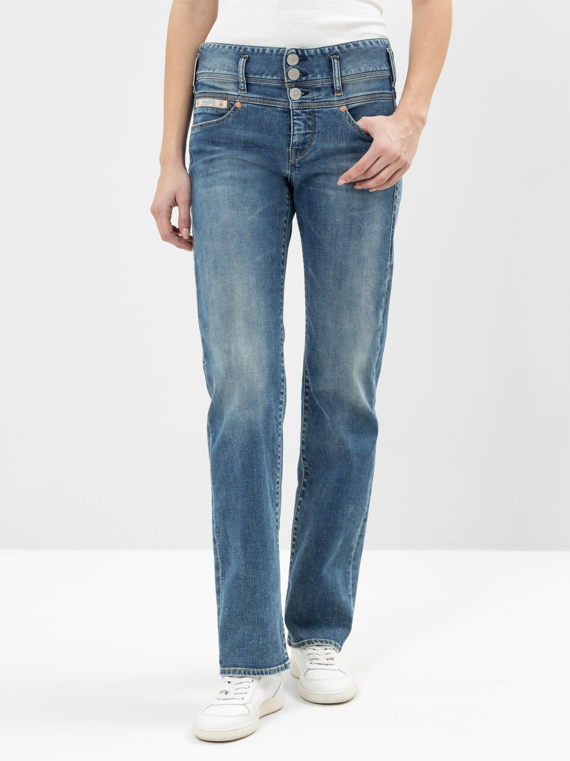 Eine Person, die hochgekrempelte blaue Jeans mit mehreren Knöpfen und weiße Turnschuhe trägt, steht vor einem schlichten hellen Hintergrund. Die Hand der Person ruht auf ihrer Hüfte.