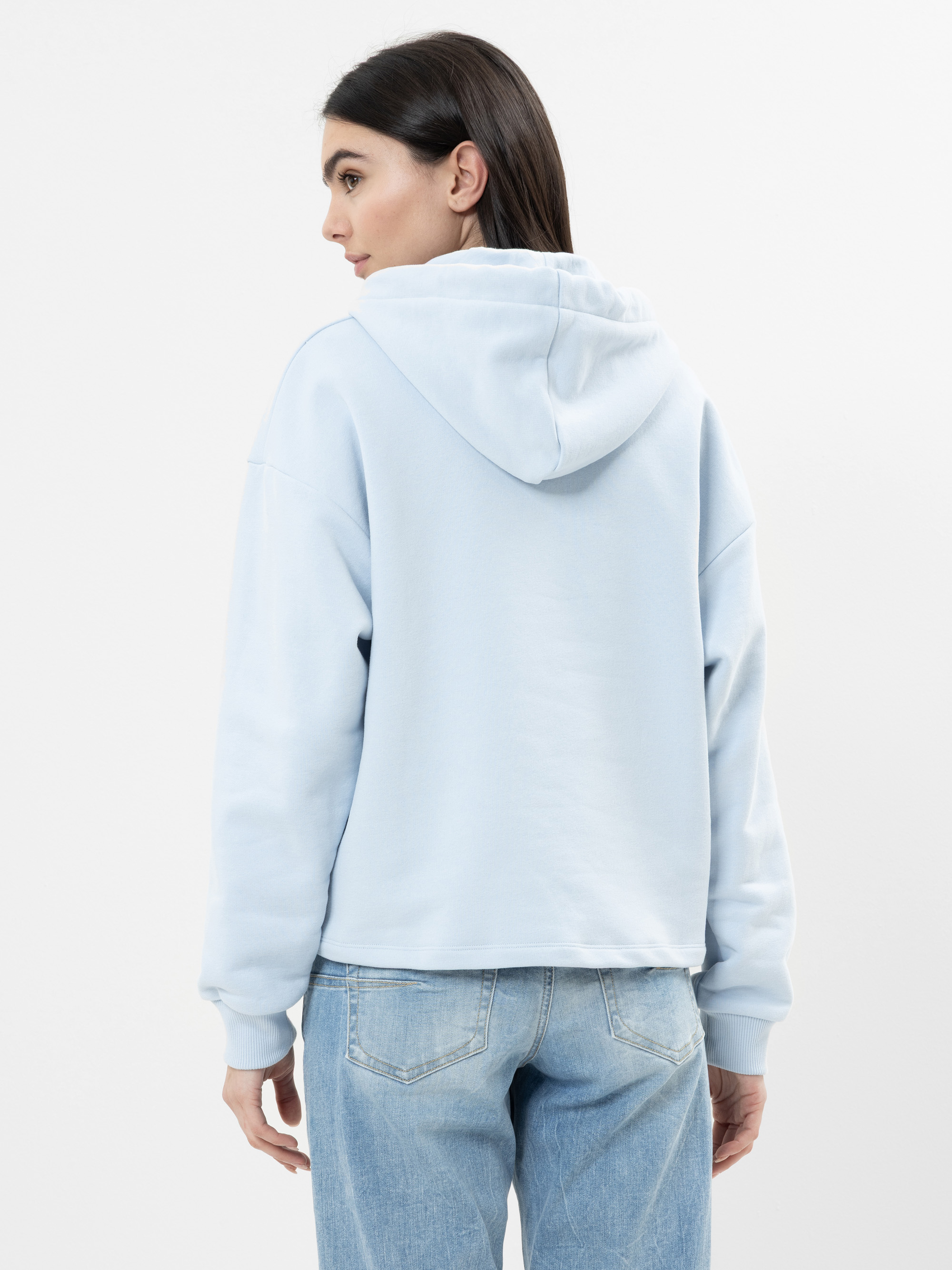 Eine Frau mit langen dunklen Haaren, bekleidet mit einem hellblauen Kapuzenpulli und hellblauen Jeans, steht mit dem Gesicht zur Wand und blickt über ihre Schulter auf einen schlichten weißen Hintergrund.